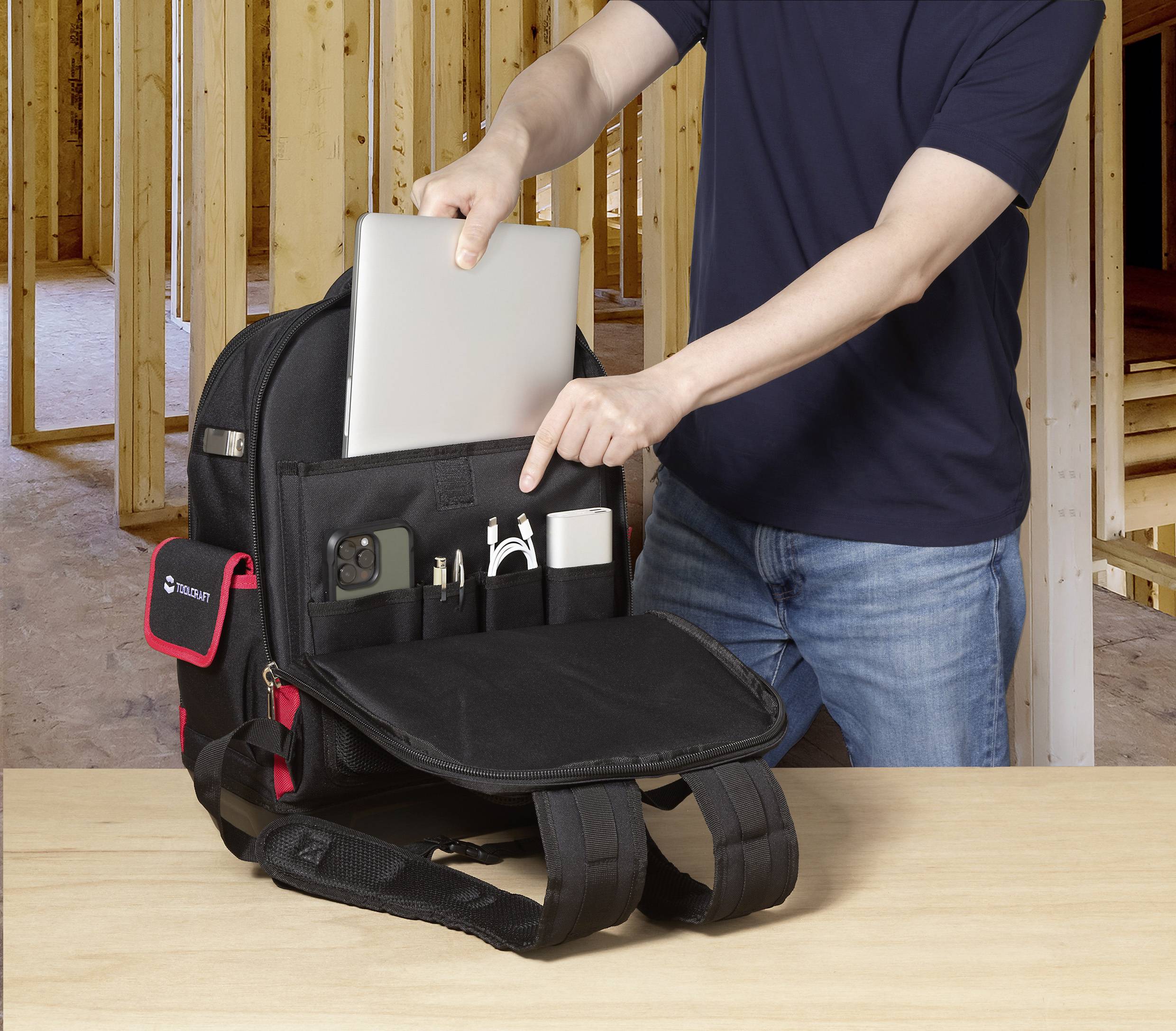 Eine Person legt ein Tablet in eine Tasche des Rucksacks, in der sich auch ein Handy, ein Ladegerät und Kabel befinden. Die Umgebung ist eine Holzbaustelle.