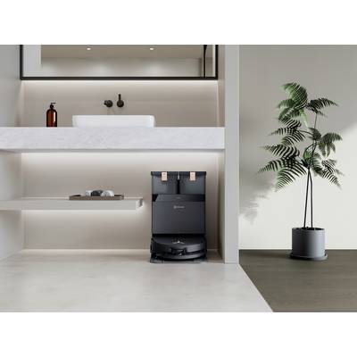 Ein Staubsauger-Roboter befindet sich in einer modernen, minimalistisch eingerichteten Wohnung neben einer Pflanze und einem Waschbecken.