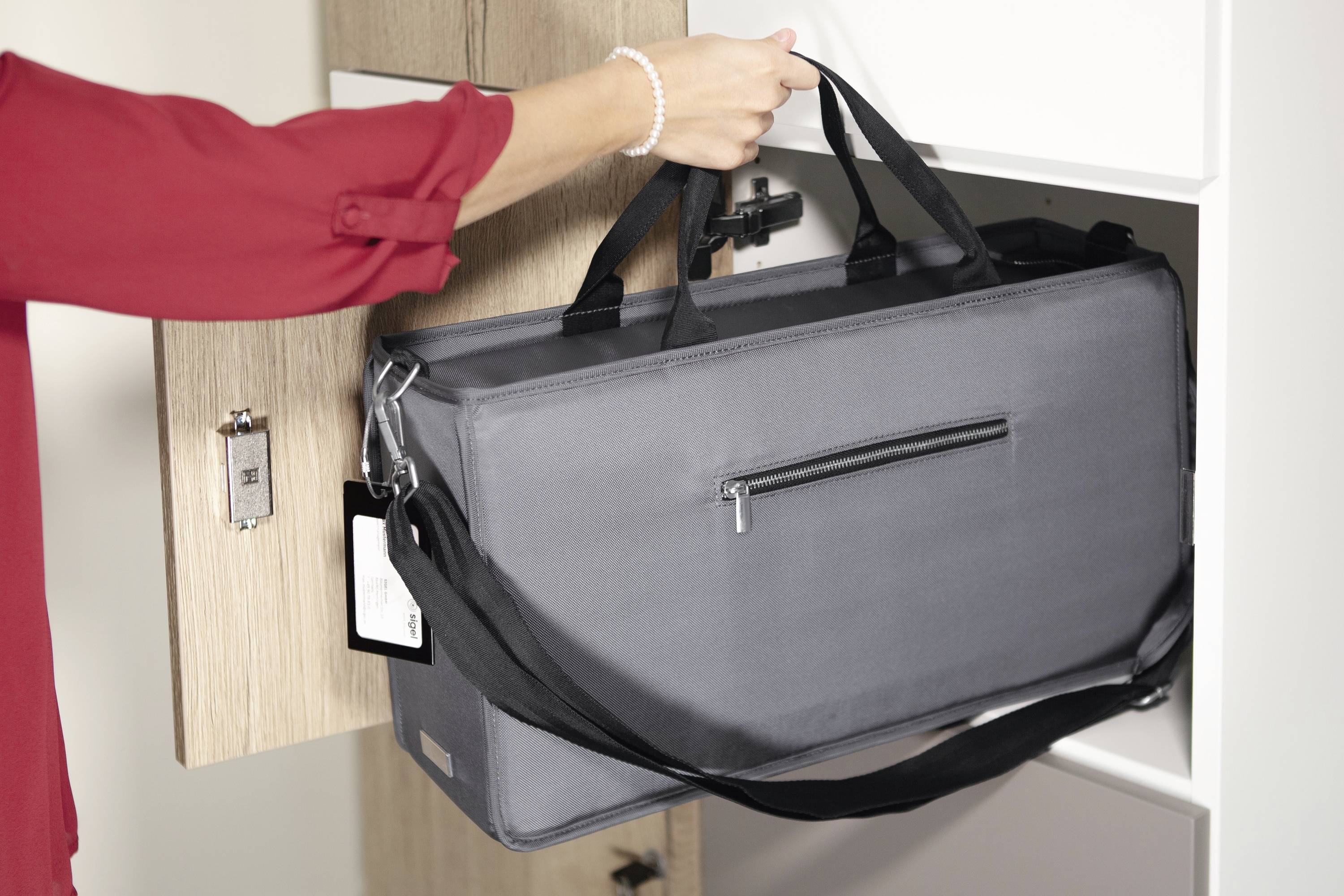 'Eine Person in roter Bluse verstaut eine graue Tragetasche in einem Schrank. Die Tasche hat Tragegriffe und einen Reißverschluss.'