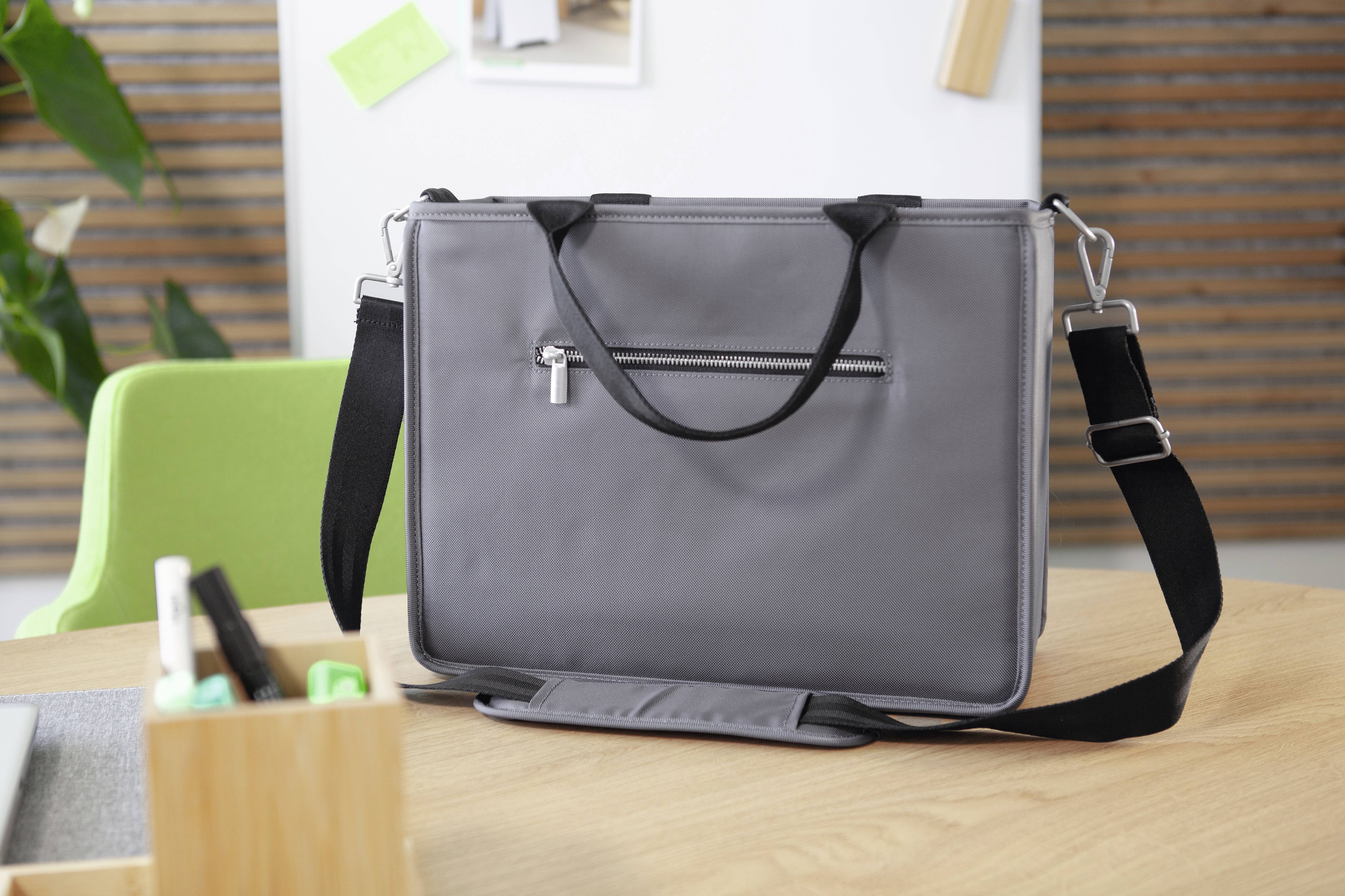 Graue Laptop-Tasche mit schwarzem Riemen und Reißverschluss, auf einem Holztisch in einem Büro mit grünem Stuhl und Pflanzen im Hintergrund.
