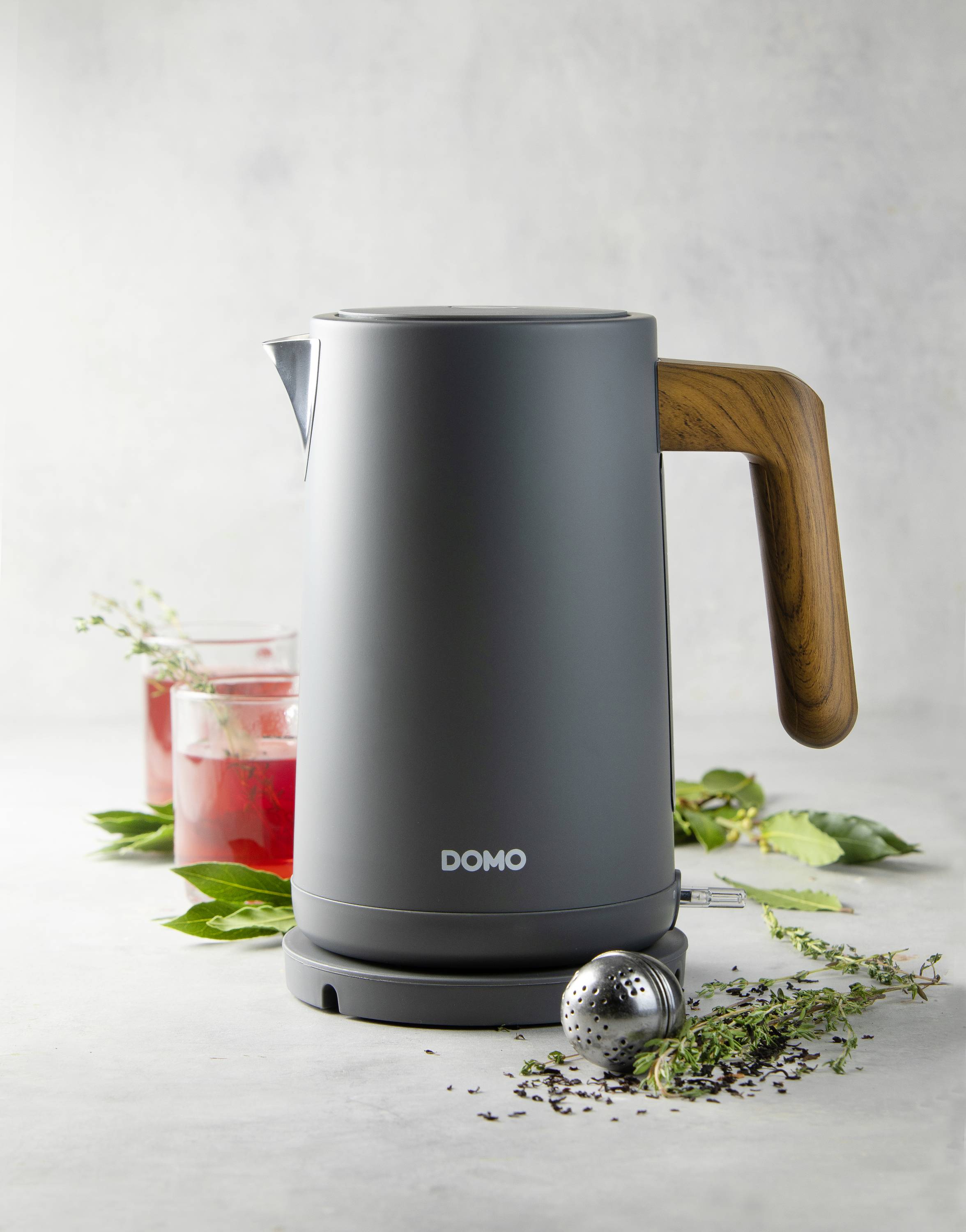 Ein grauer Wasserkocher mit Holzgriff steht auf einem Tisch neben einem Glas mit rotem Getränk und Kräutern. Markierung: 'DOMO'.