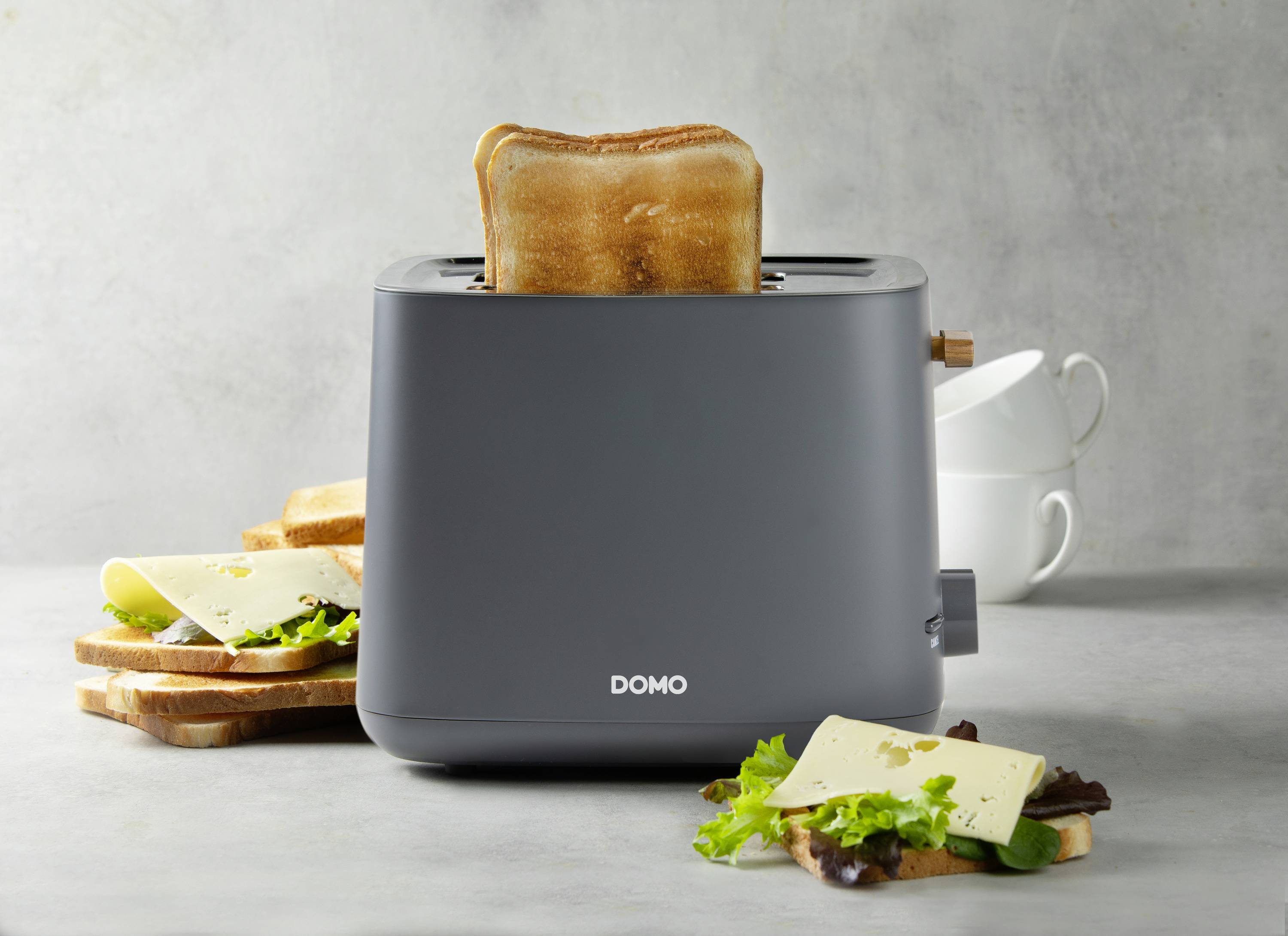 Ein grauer Toaster mit der Marke 'DOMO' toastet eine Scheibe Brot. Daneben liegen belegte Brote mit Salat und Käse.
