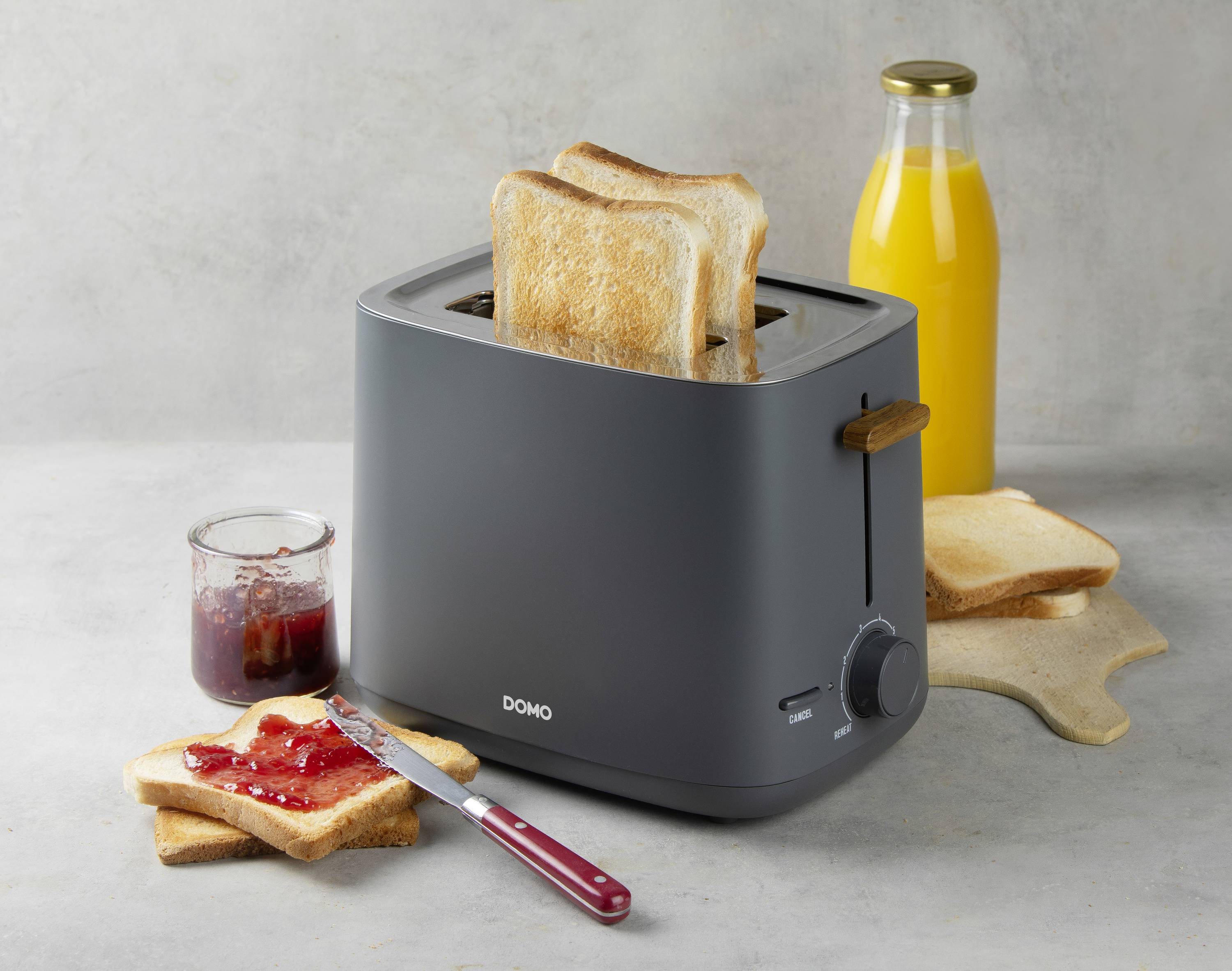 Ein schwarzer Toaster mit zwei Scheiben goldbraunem Toast. Daneben stehen ein Glas Orangennektar, ein Marmeladenglas und ein Messer auf Toast.