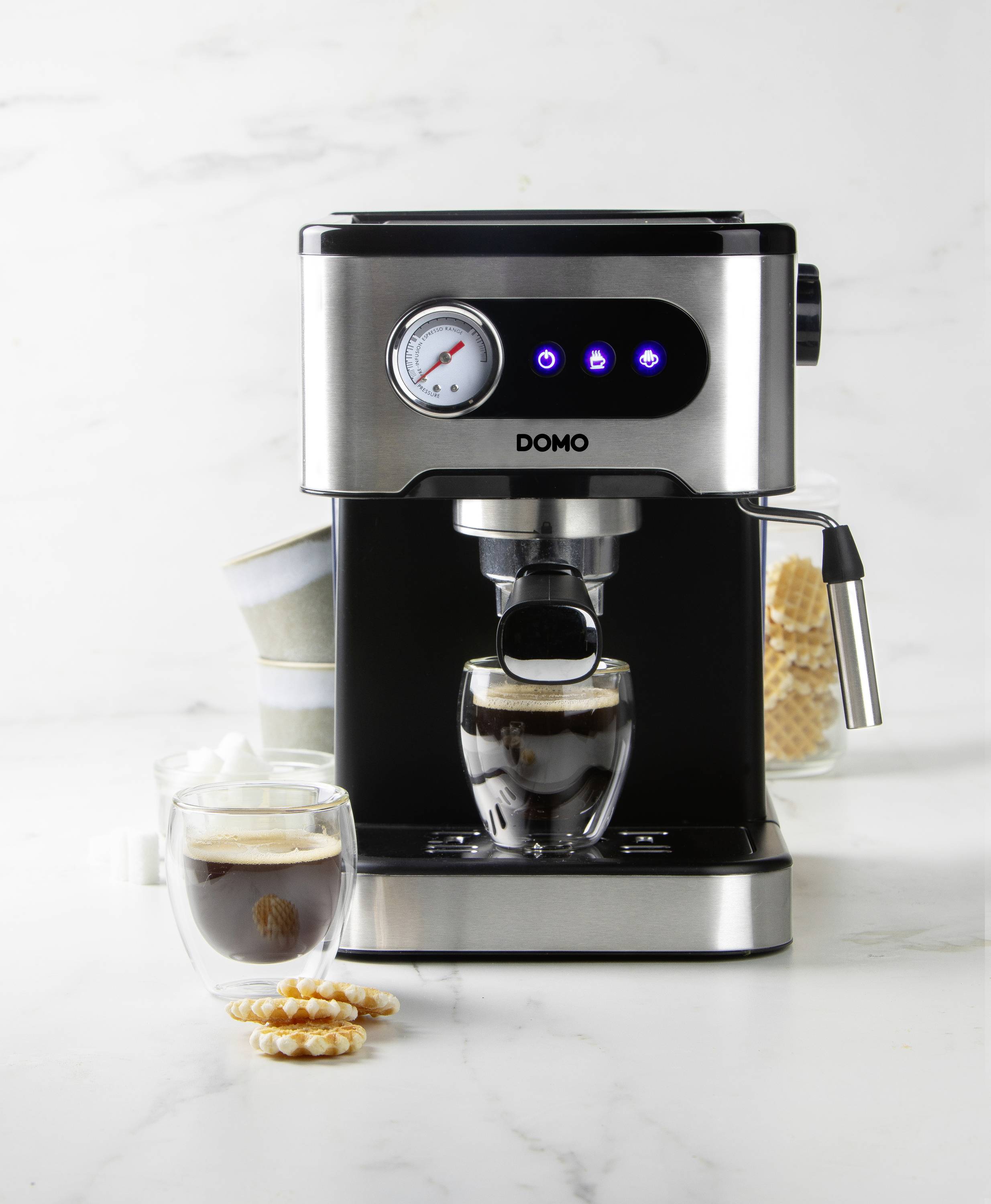 Espressomaschine mit zwei Gläsern Espresso davor, gestapelte Tassen und Zucker im Hintergrund; moderne, elegante Küchengeräte umgeben.