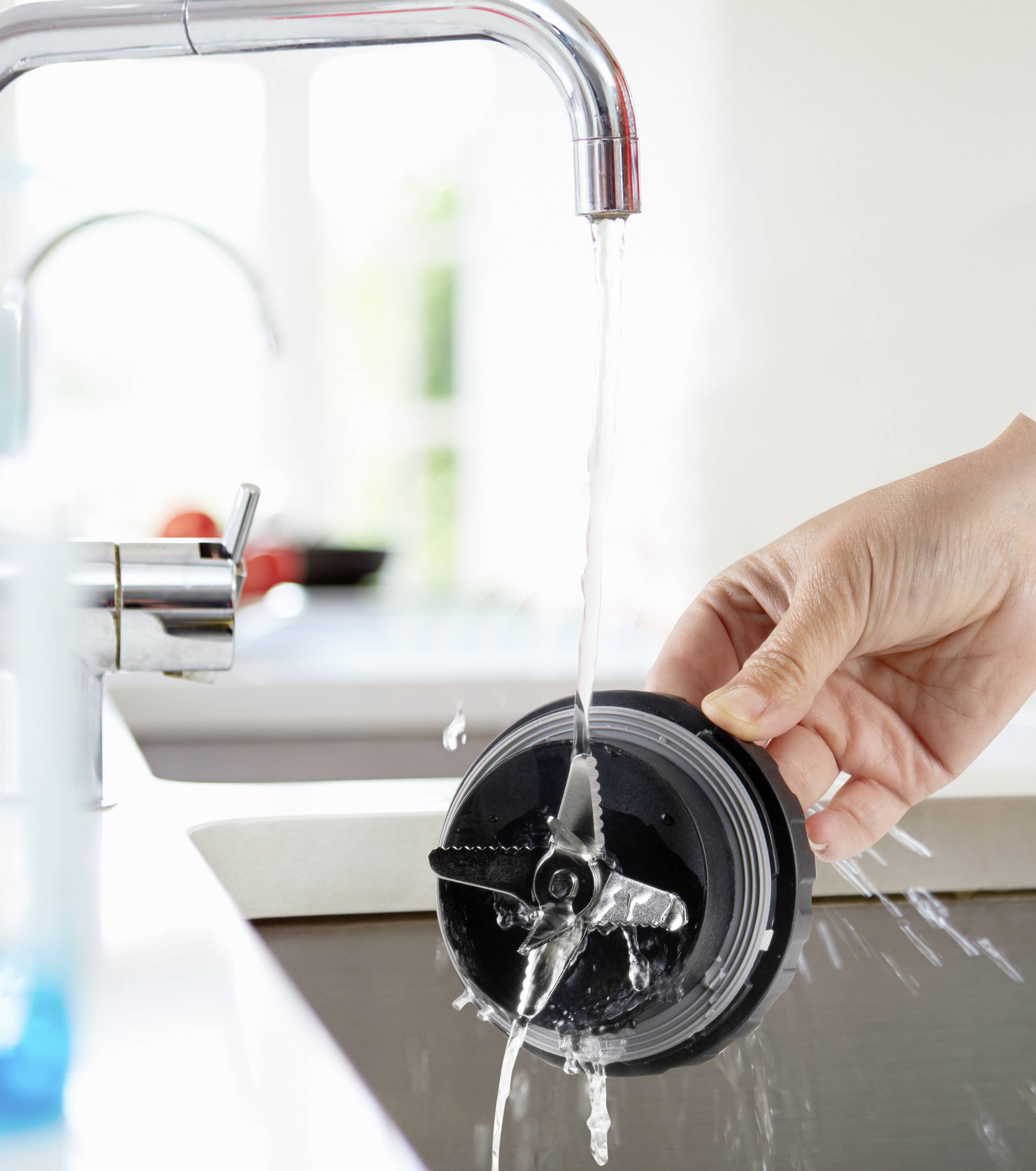 Eine Hand wäscht einen Mixeraufsatz unter fließendem Wasser in einem Waschbecken. Hintergrund zeigt eine helle Küche.