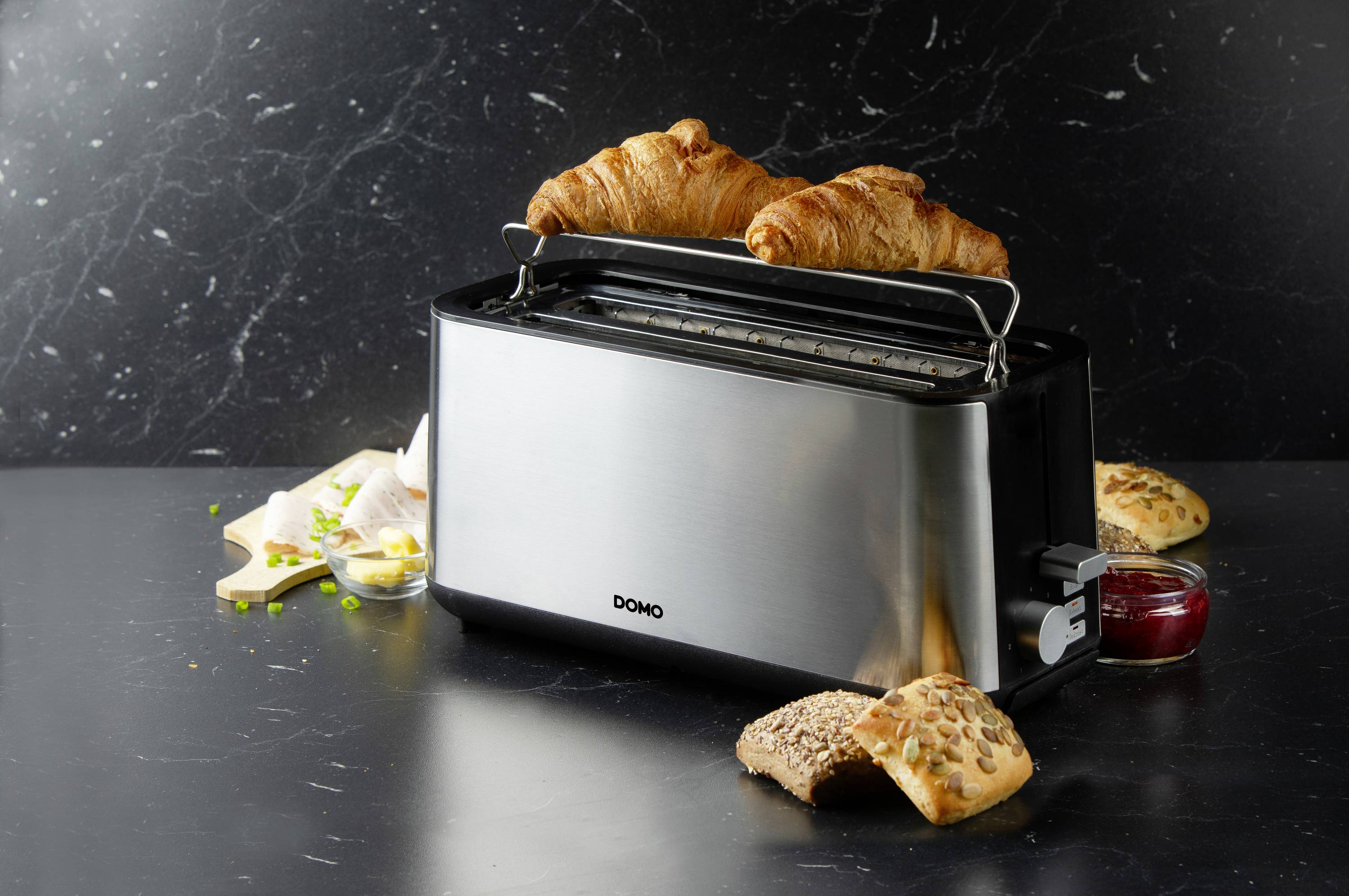 Ein silberner Toaster mit zwei Croissants oben drauf. Daneben liegen frisches Brot, Marmelade und ein Messer auf einem dunklen Tisch.