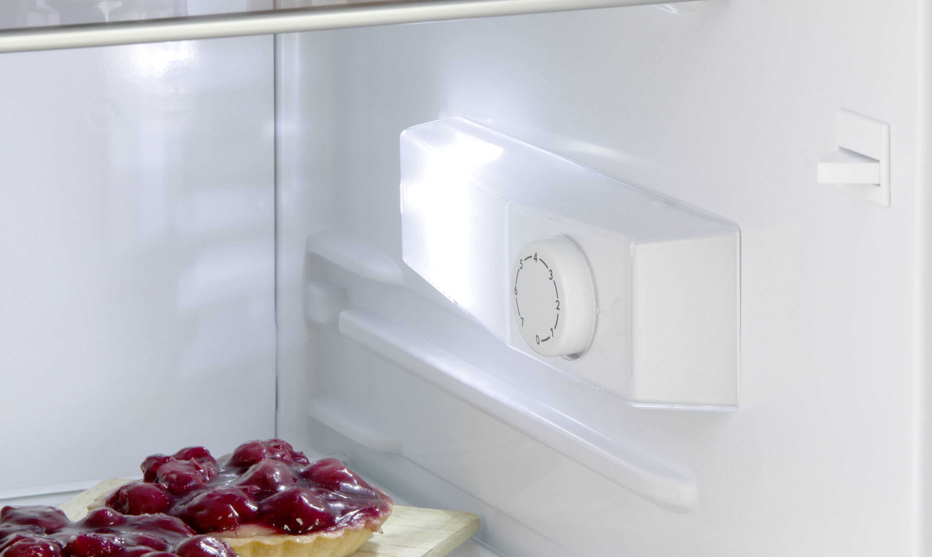 Ein Kuchen mit Kirschen auf einem Holzbrett steht in einem beleuchteten und leeren Kühlschrank.