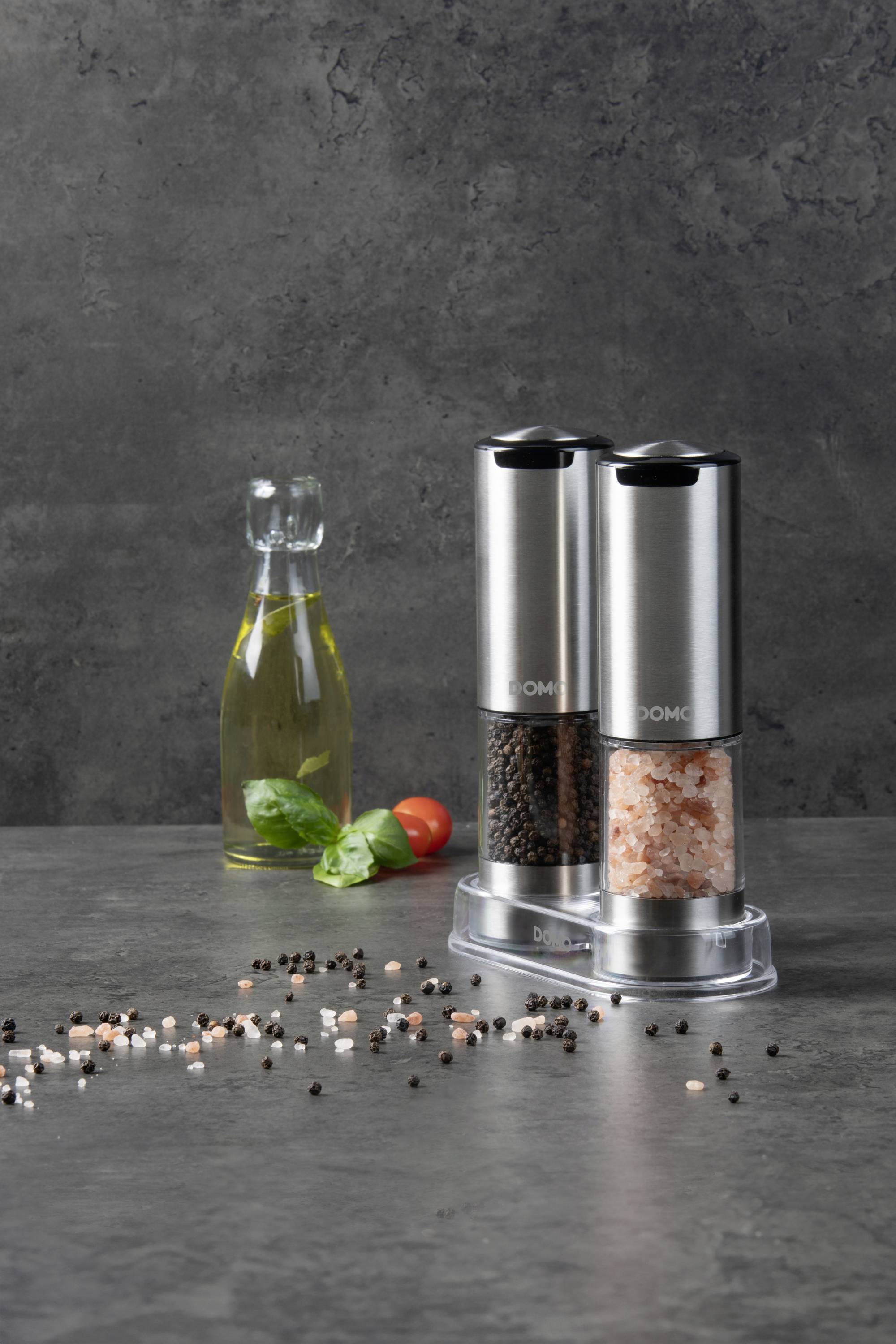 Zwei Edelstahl-Gewürzmühlen mit schwarzem Pfeffer und rosa Salz stehen auf einem Tisch. Daneben eine Ölflasche und Basilikumblätter.