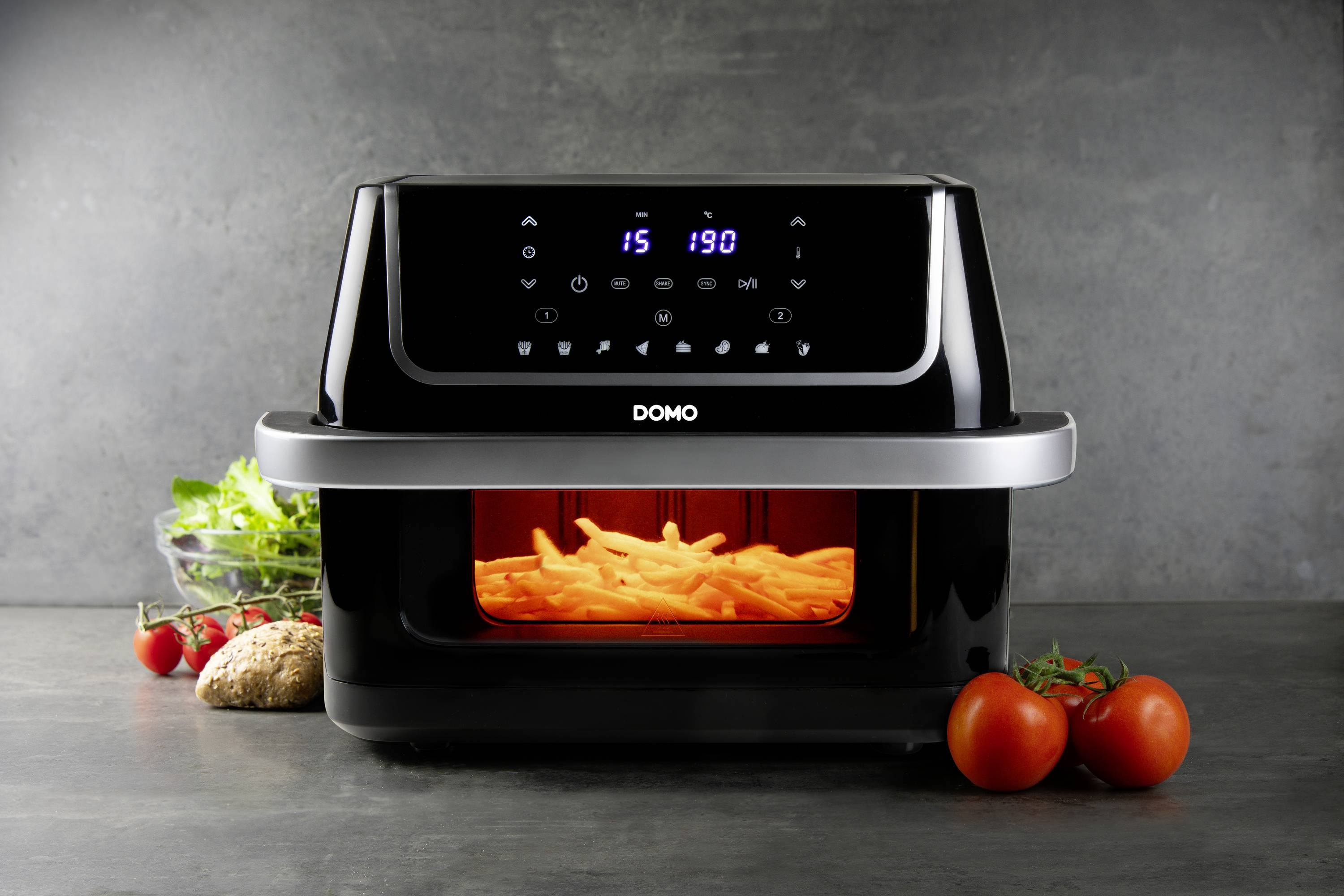 'Schwarzer Ofen mit Touchscreen, backt Pommes. Im Vordergrund frische Tomaten und Salat. Moderne Küchenszene.'