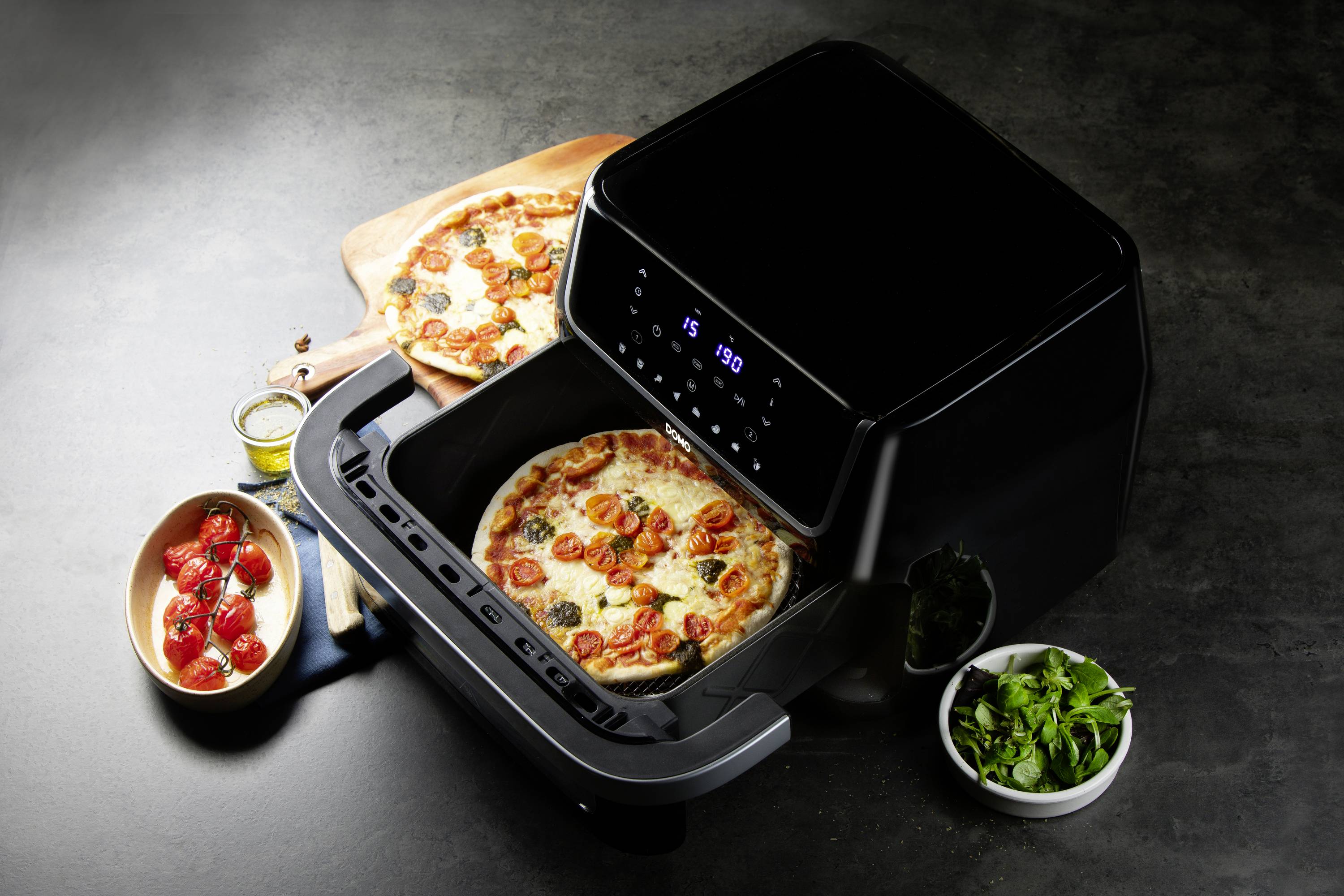 Eine Pizza liegt in einer Heißluftfritteuse auf einem Tisch. Daneben stehen frische Zutaten und eine Schüssel mit Kräutern.