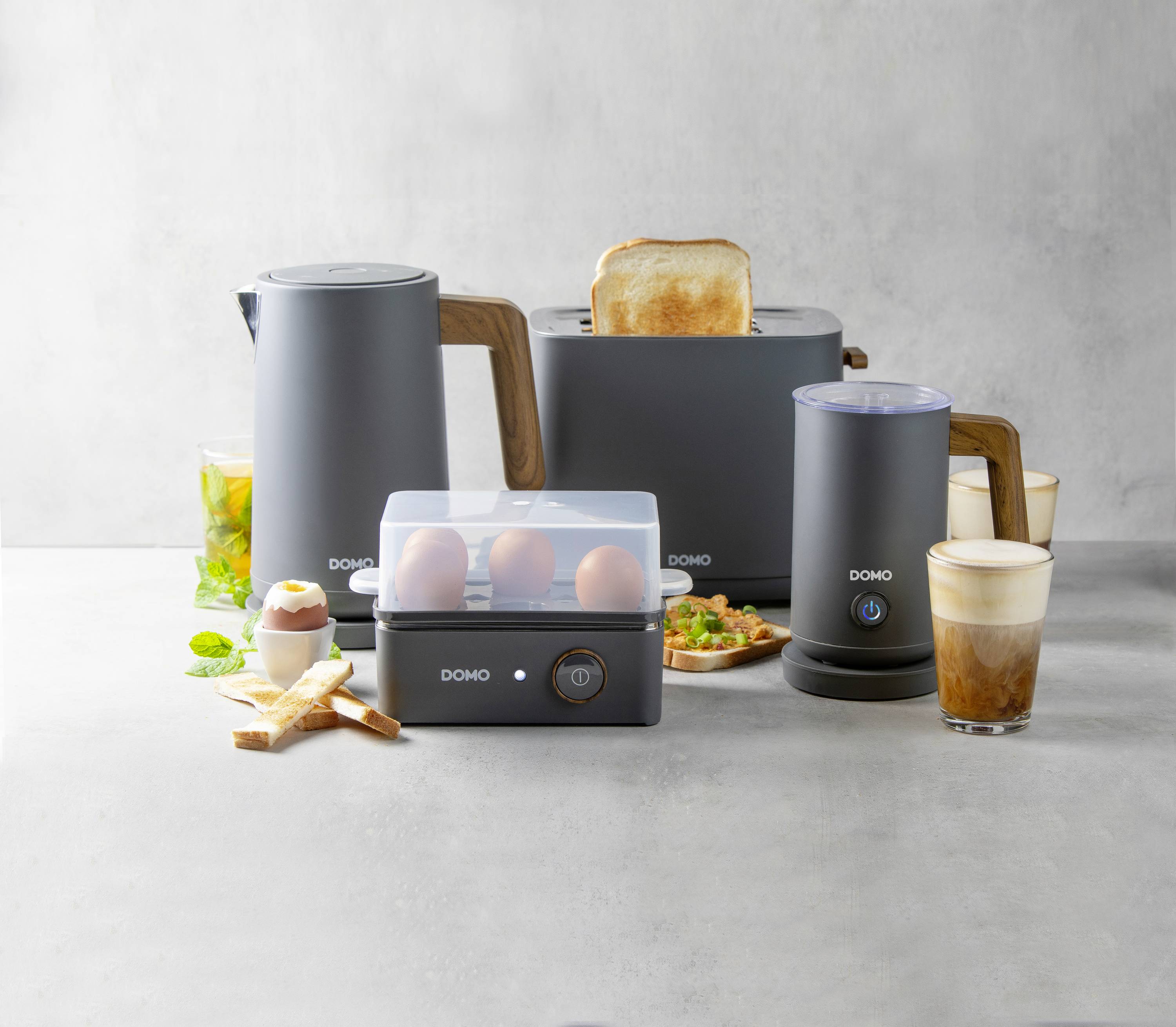 Eine graue Küchengerätekollektion, bestehend aus Wasserkocher, Toaster mit Toastscheibe, Eierkocher und Milchaufschäumer, auf grauem Hintergrund.