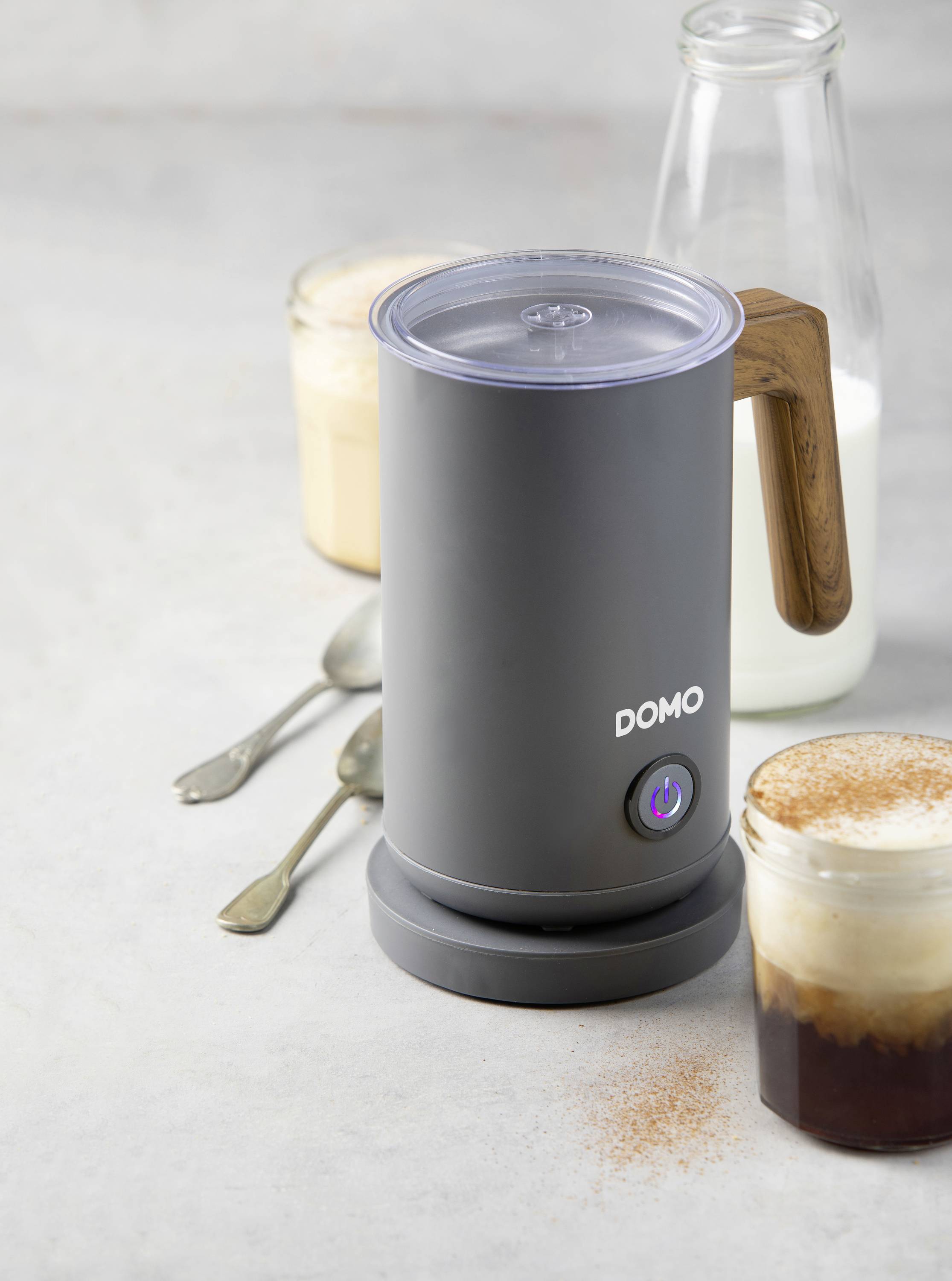 Ein elektrischer Milchaufschäumer mit Holzgriff steht auf einem Tisch neben zwei Gläsern Milchkaffee und einem Glas Milch.