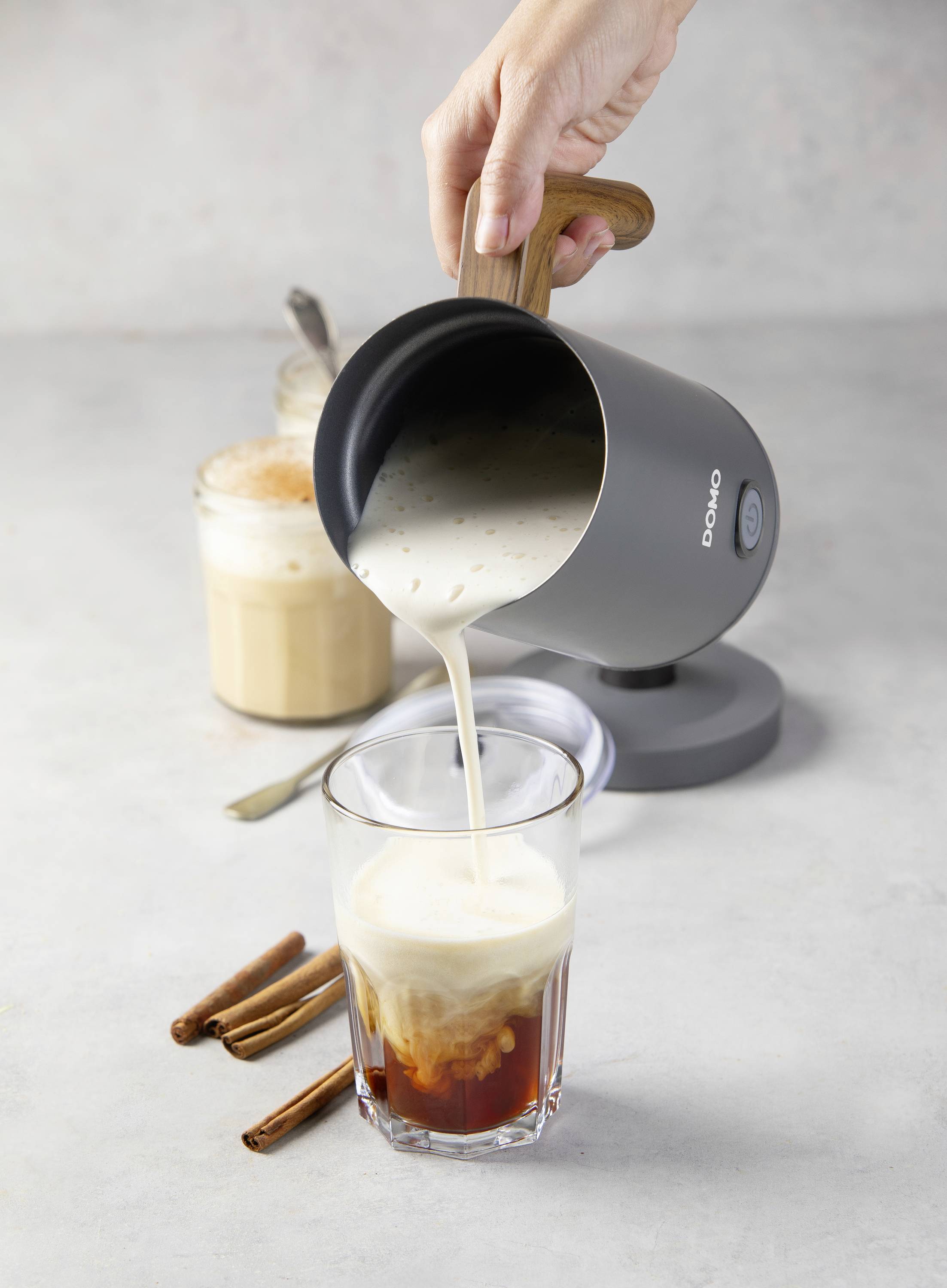 Eine Hand gießt Milch aus einem elektrischen Milchaufschäumer in ein Glas, daneben Kaffee und Zimtstangen auf einem Tisch.