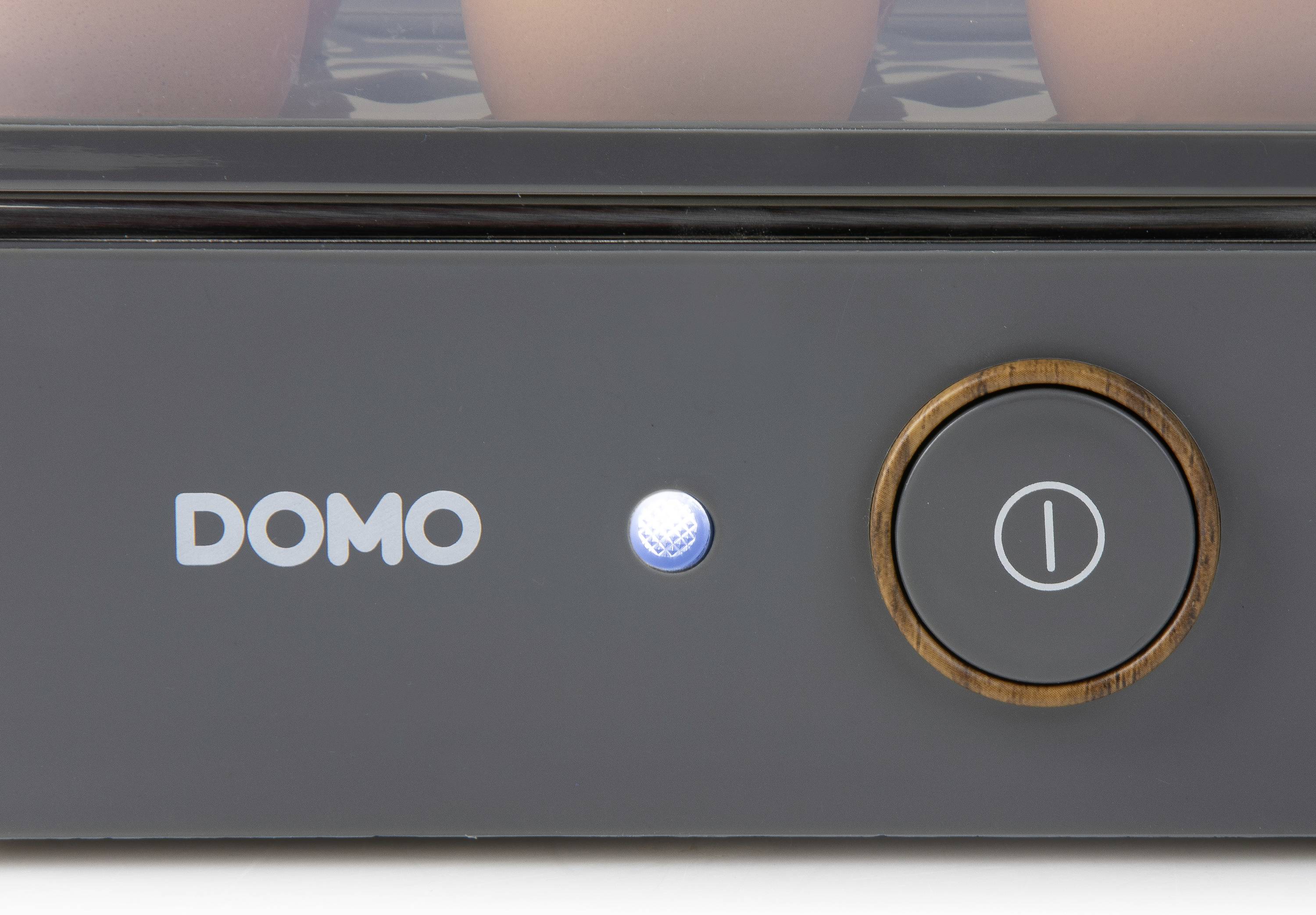 Ein Ausschnitt eines Elektrogeräts zeigt einen 'DOMO'-Schriftzug und einen runden Ein-/Ausschaltknopf mit einem blauen Lämpchen daneben.