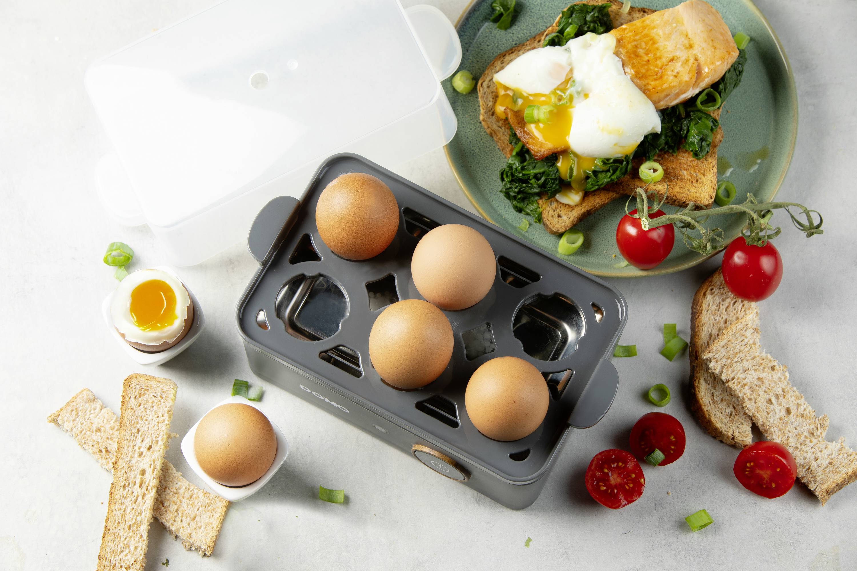 Frühstückszene mit einem Gerät zum Eierkochen, fünf Eiern, einem aufgeschnittenen Ei, Toaststreifen, Spinat und pochierten Eiern auf Brot.