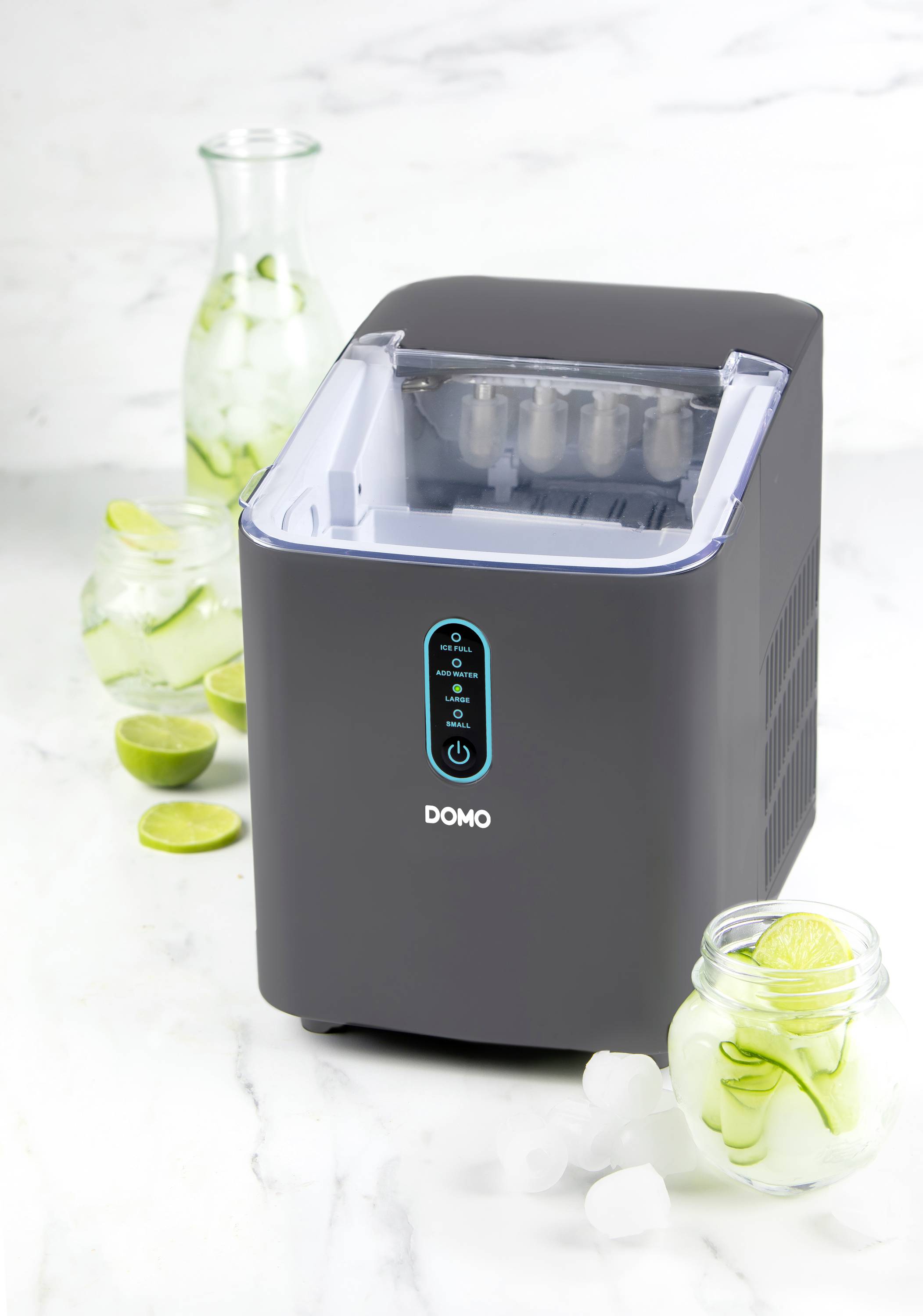 Ein grauer Eiswürfelbereiter steht auf einem Tisch. Daneben Gläser mit Limettenscheiben und Eis sowie eine Glaskaraffe.