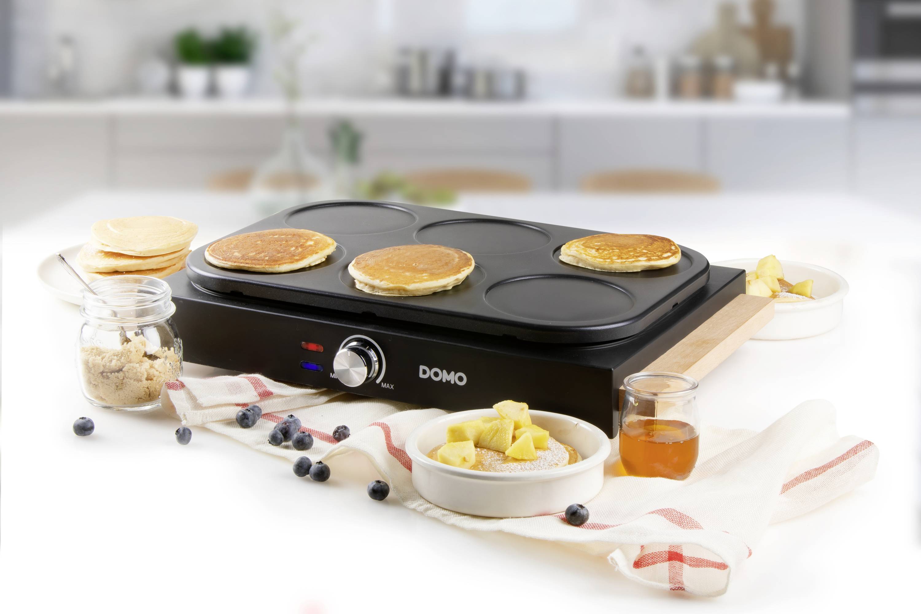 Ein Elektrogrill mit vier Pfannkuchen darauf, umgeben von Zutaten wie Ananas, Honig und Blaubeeren in einer modernen Küche.