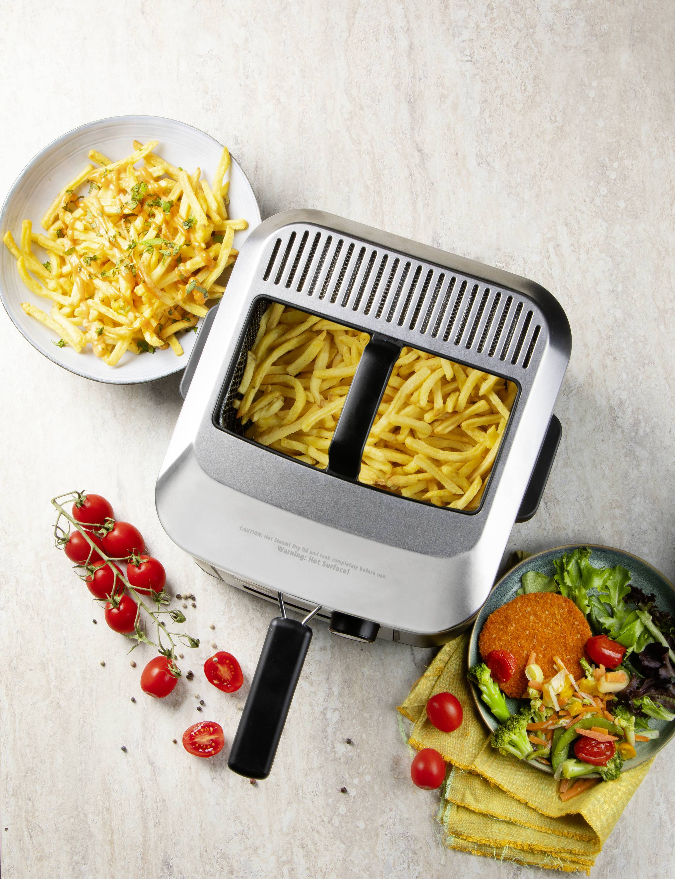 Eine Heißluftfritteuse mit zubereiteten Pommes auf einem Tisch. Daneben stehen zwei Schalen: eine mit Salat, die andere mit Pommes.