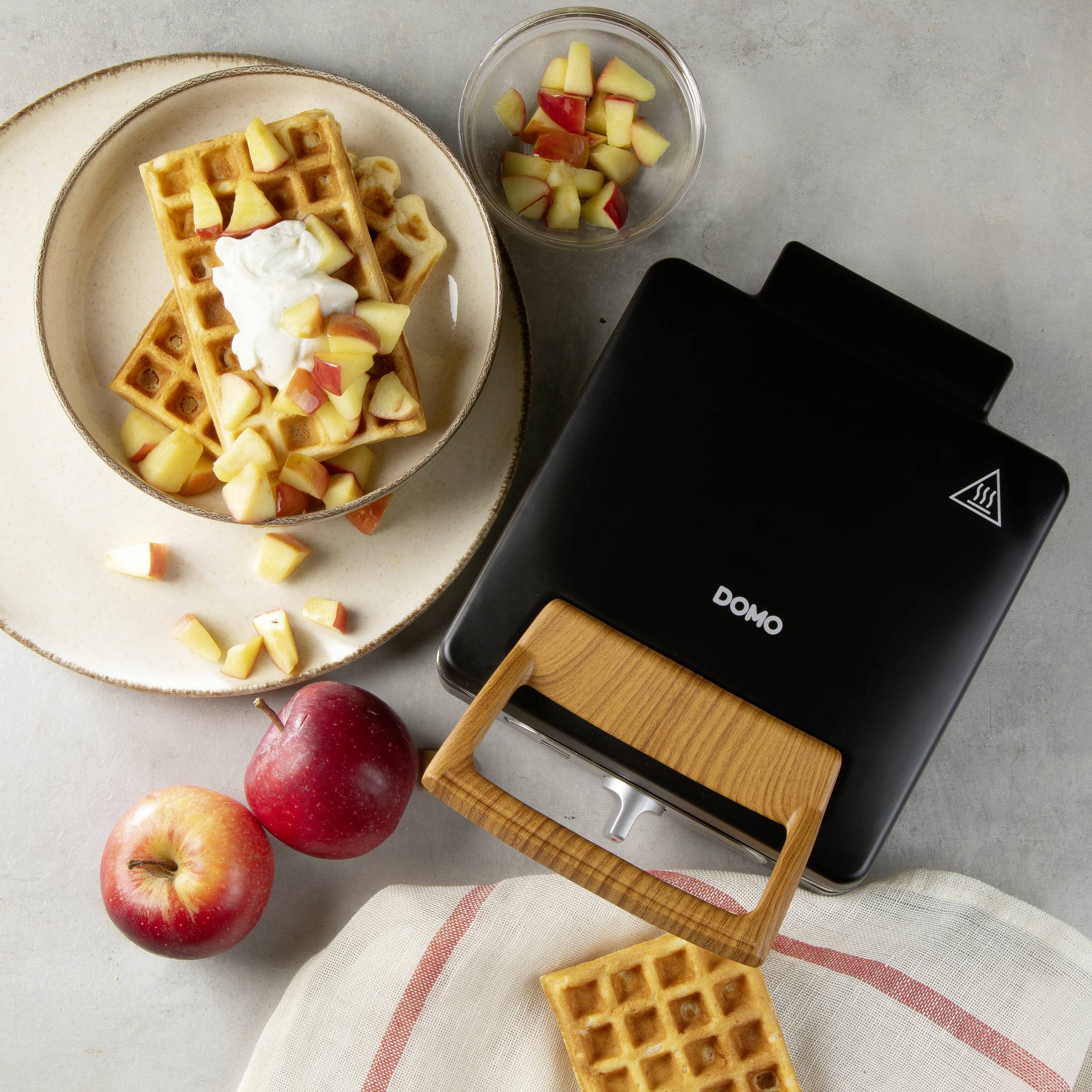 Waffeleisen mit Waffel auf Holztisch, plus Teller mit zwei Waffeln, Apfelstücken und Sahne. Zwei ganze Äpfel und extra Apfelstücke in Schüssel.