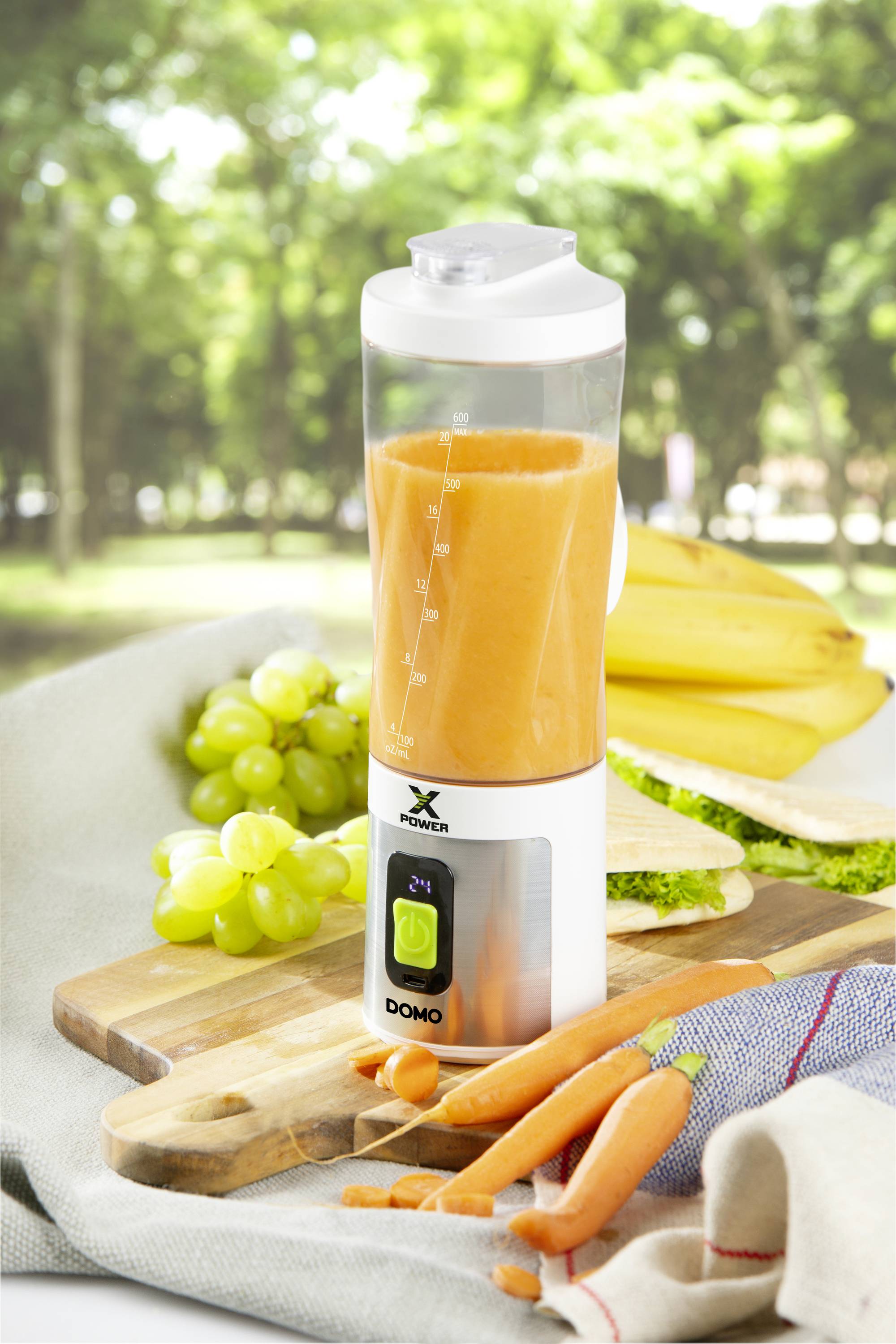 Ein Mixer gefüllt mit orangem Saft steht auf einem Schneidebrett neben Weintrauben, Karotten und einem Sandwich im Freien.