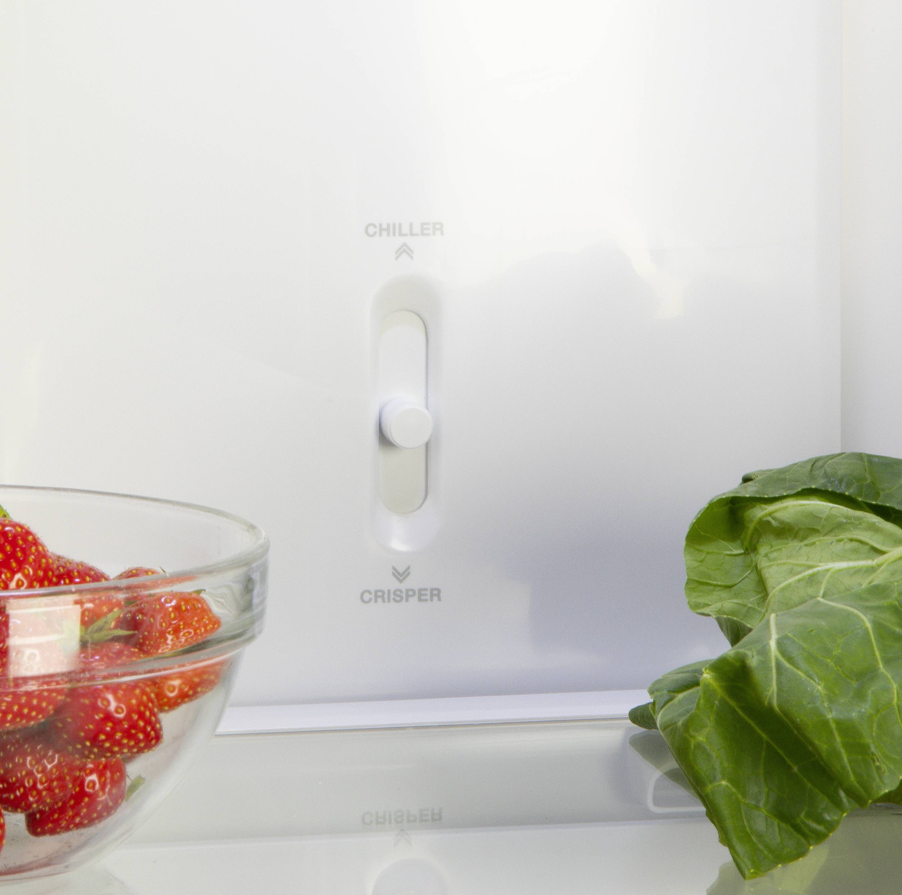 Eine Glasschüssel mit Erdbeeren steht neben grünem Blattgemüse vor einem Kühlschrank, der einen Regler für 'Chiller' und 'Crisper' zeigt.
