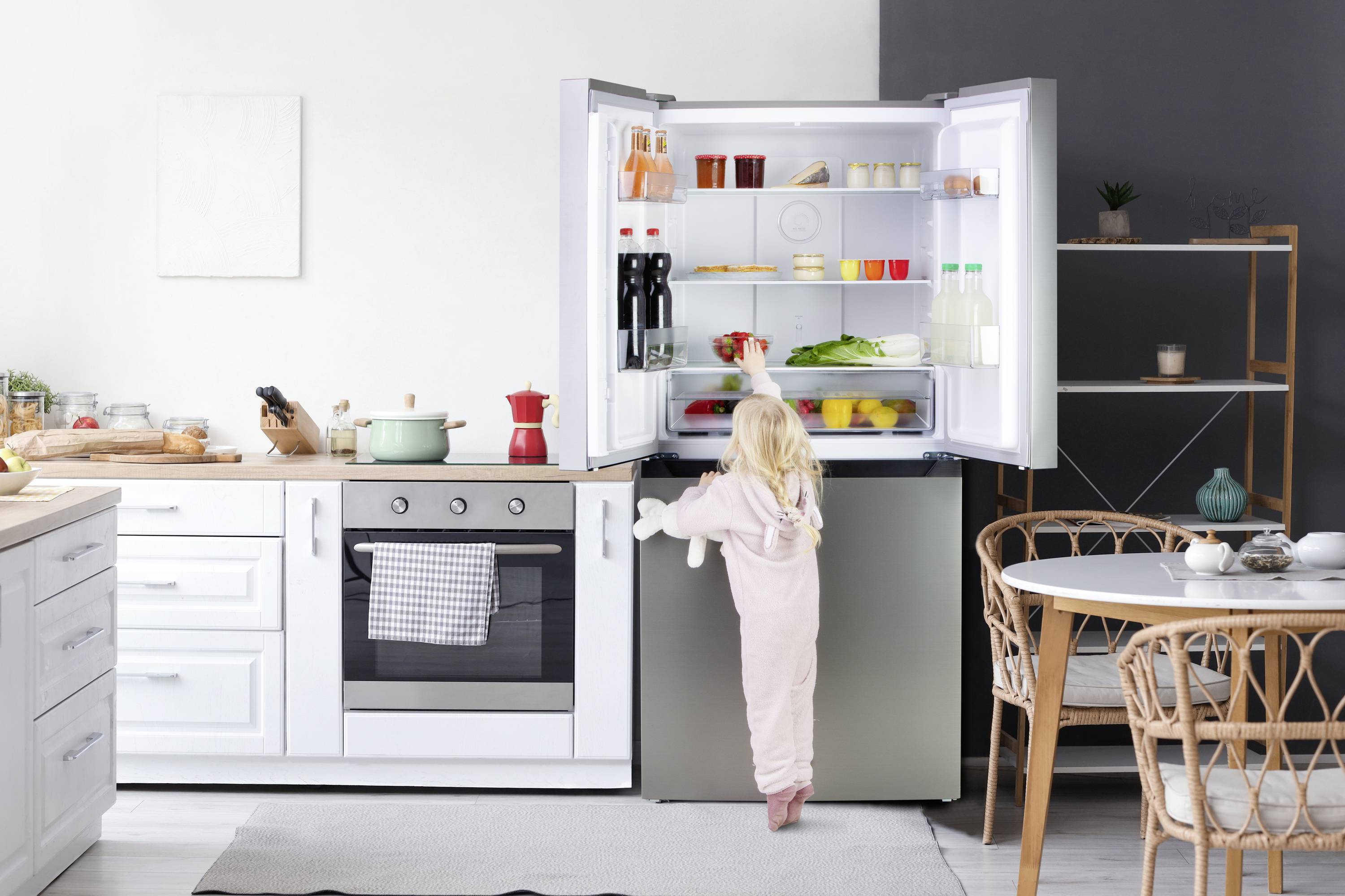 Ein kleines Mädchen steht in der Küche und schaut in einen offenen Kühlschrank, der mit Lebensmitteln gefüllt ist.