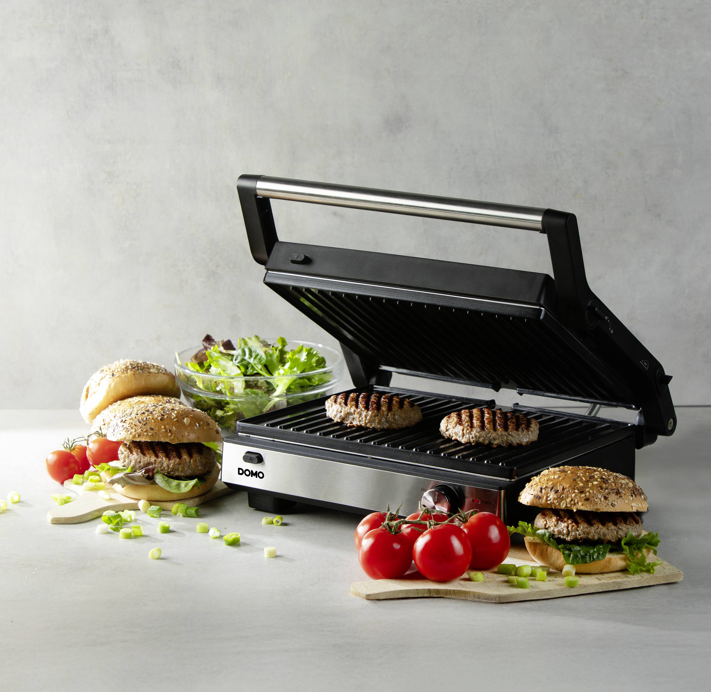 Ein offener Kontaktgrill mit zwei Burger-Patties darauf. Daneben liegen fertige Hamburger mit Salat und Tomaten als Beilagen.