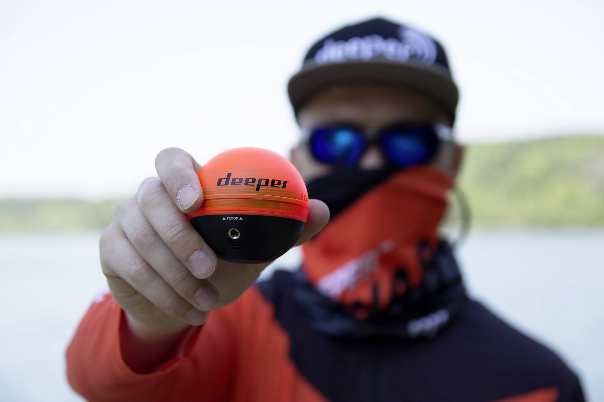 Ein Angler hält einen orange-schwarzen Fischfinder mit der Aufschrift 'deeper' in der Hand. Hintergrund unscharf mit Wasser und Bäumen.