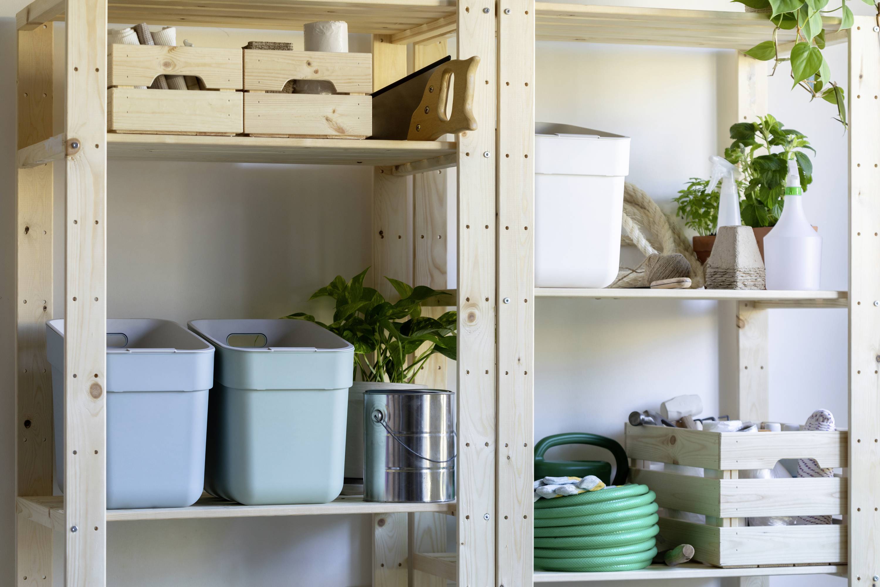 Holzregale in einer Garage mit blauen Behältern, Pflanzen, einem Eimer und einem grünen Gartenschlauch. Ordentlich und funktional organisiert.