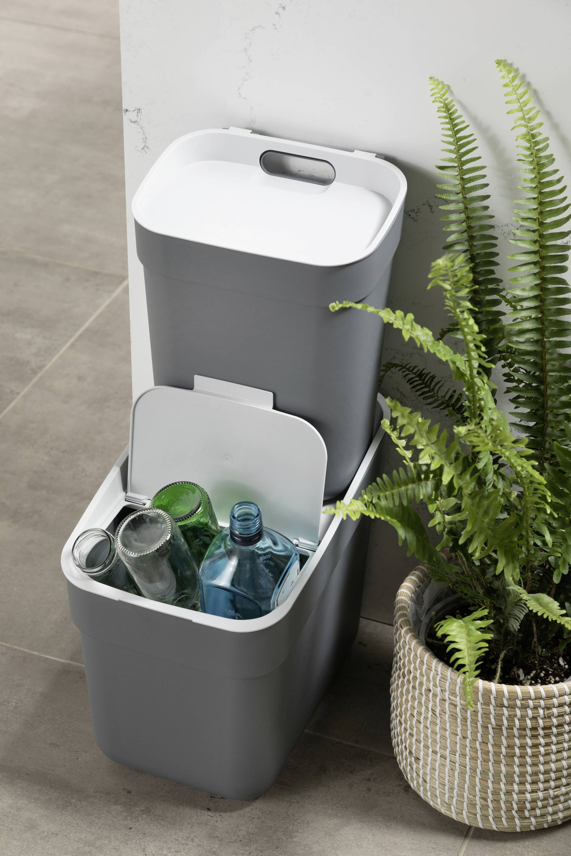 Zwei gestapelte Recyclingbehälter mit weißen Deckeln für Flaschen, daneben steht ein Farn in einem geflochtenen Topf.