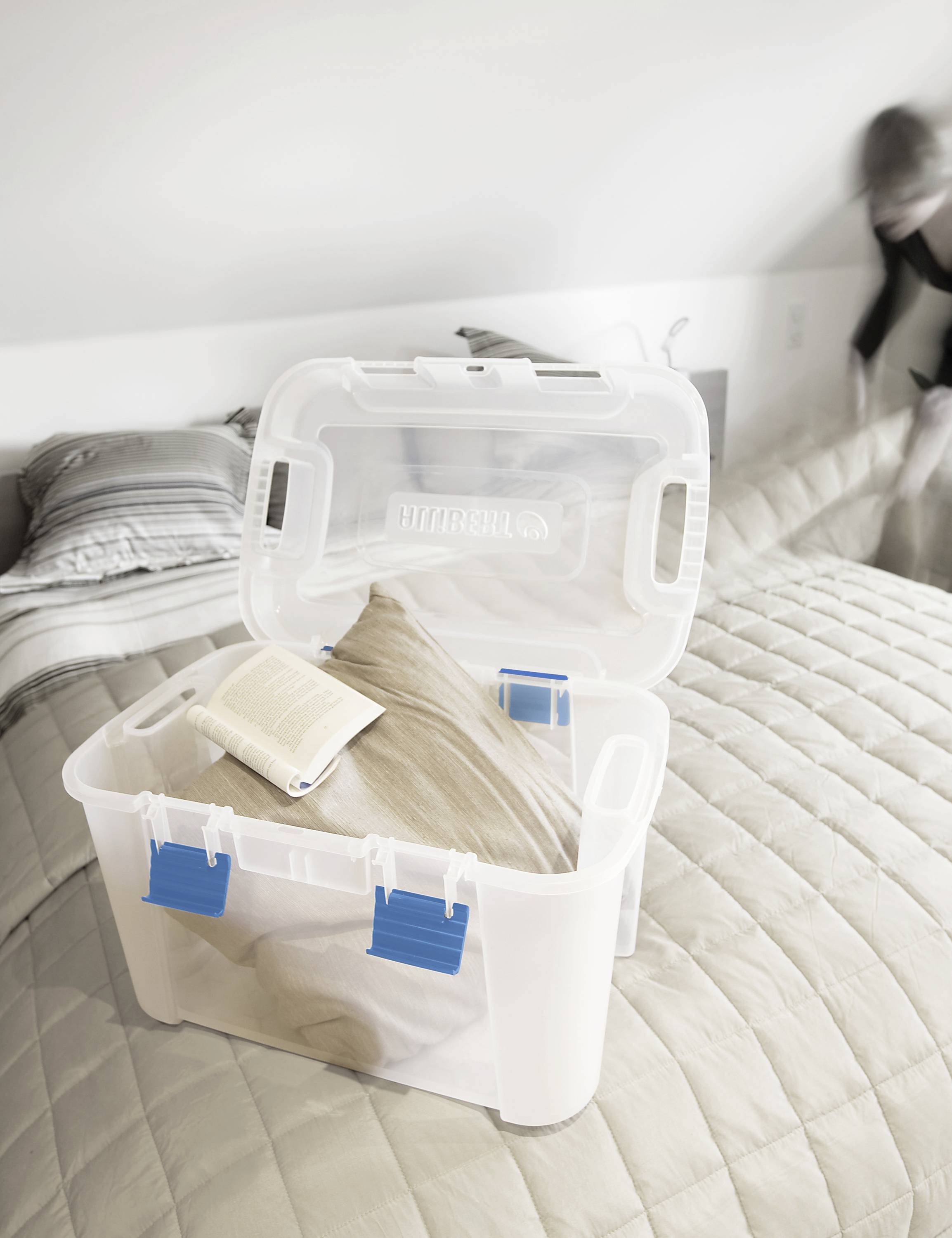 Ein geöffneter Plastikbehälter auf einem Bett enthält ein Kissen und ein Buch. Im Hintergrund richtet jemand das Bett her.