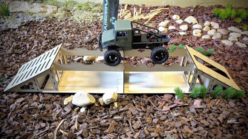 Ein Modellauto fährt über eine kleine, detailreiche Metallbrücke in einem Miniatur-Gelände mit Kieselsteinen und Pflanzen im Hintergrund.