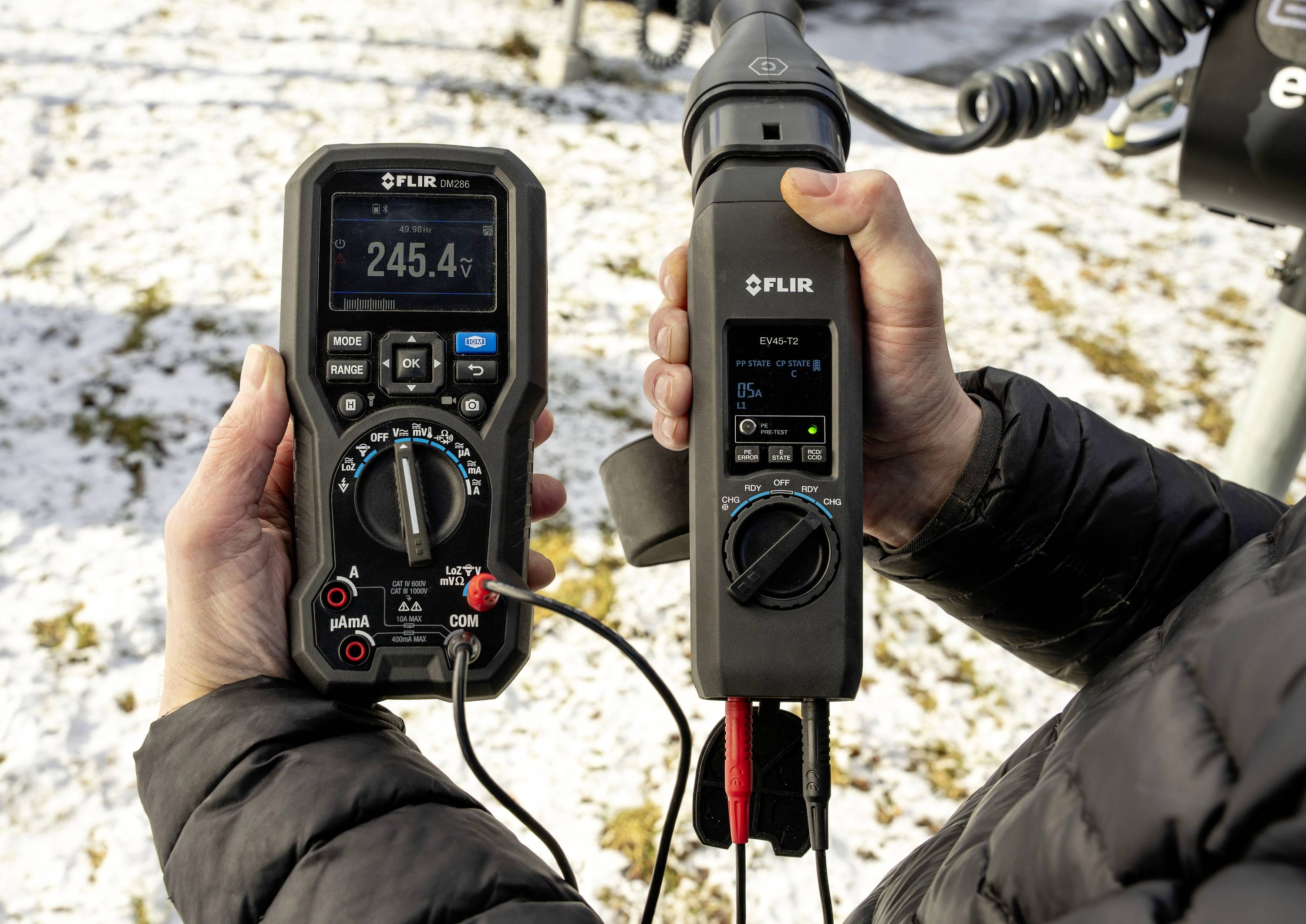 'Person hält FLIR-Messgeräte im Freien. Das linke Display zeigt 245,4V, das rechte zeigt 0,0 Lux.'