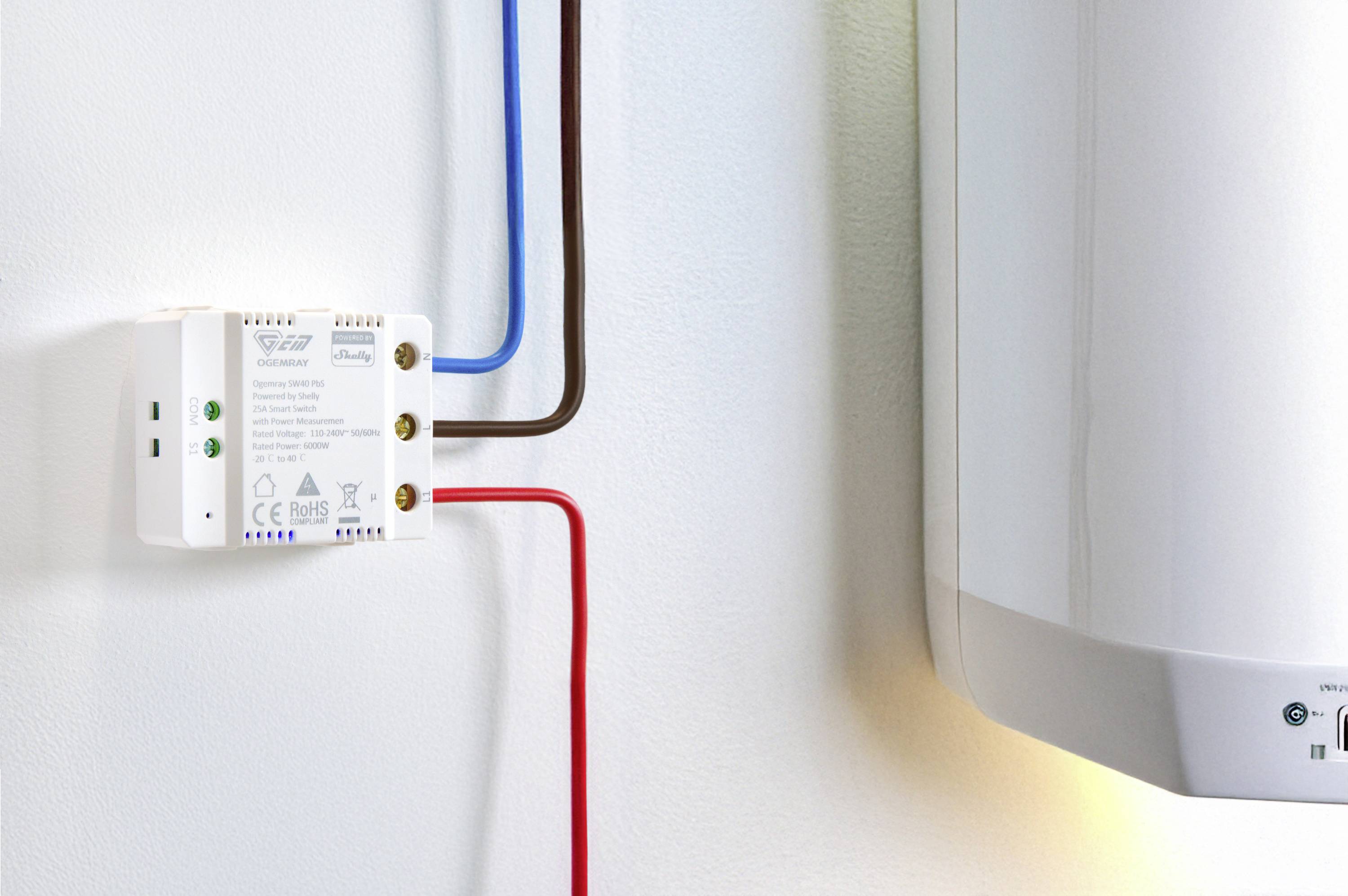 Ein weißes elektrisches Gerät mit grünen, blauen und roten Kabeln ist an einer weißen Wand montiert. Daneben befindet sich ein großer, weißer Heizboiler.