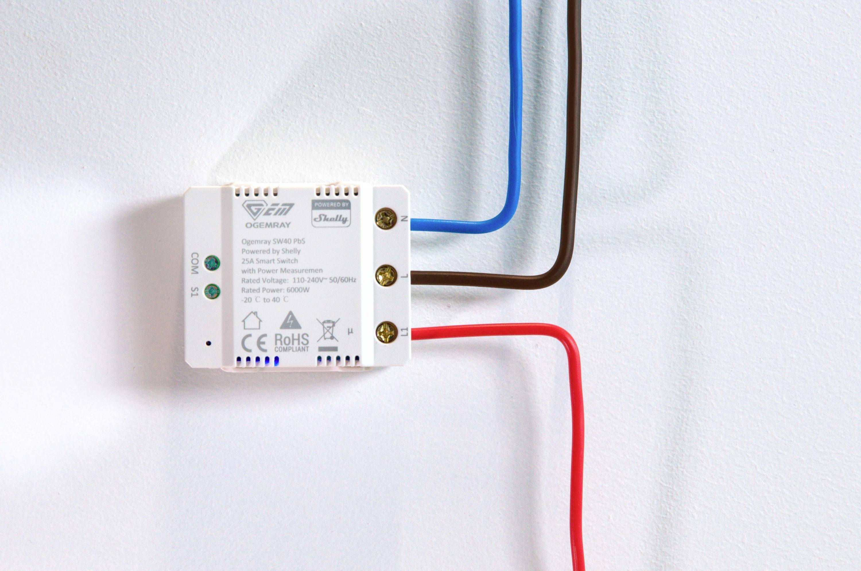 Kleines elektrisches Gerät an einer weißen Wand befestigt, mit drei angeschlossenen Drähten in blau, braun und rot.