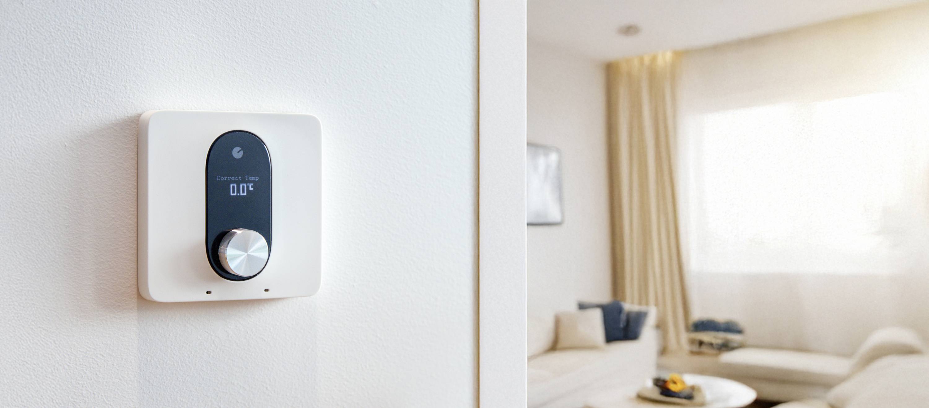 Ein Thermostat an der Wand zeigt die Temperatur an. Im Hintergrund ist ein heller Wohnbereich mit Sofa und Fenster zu sehen.