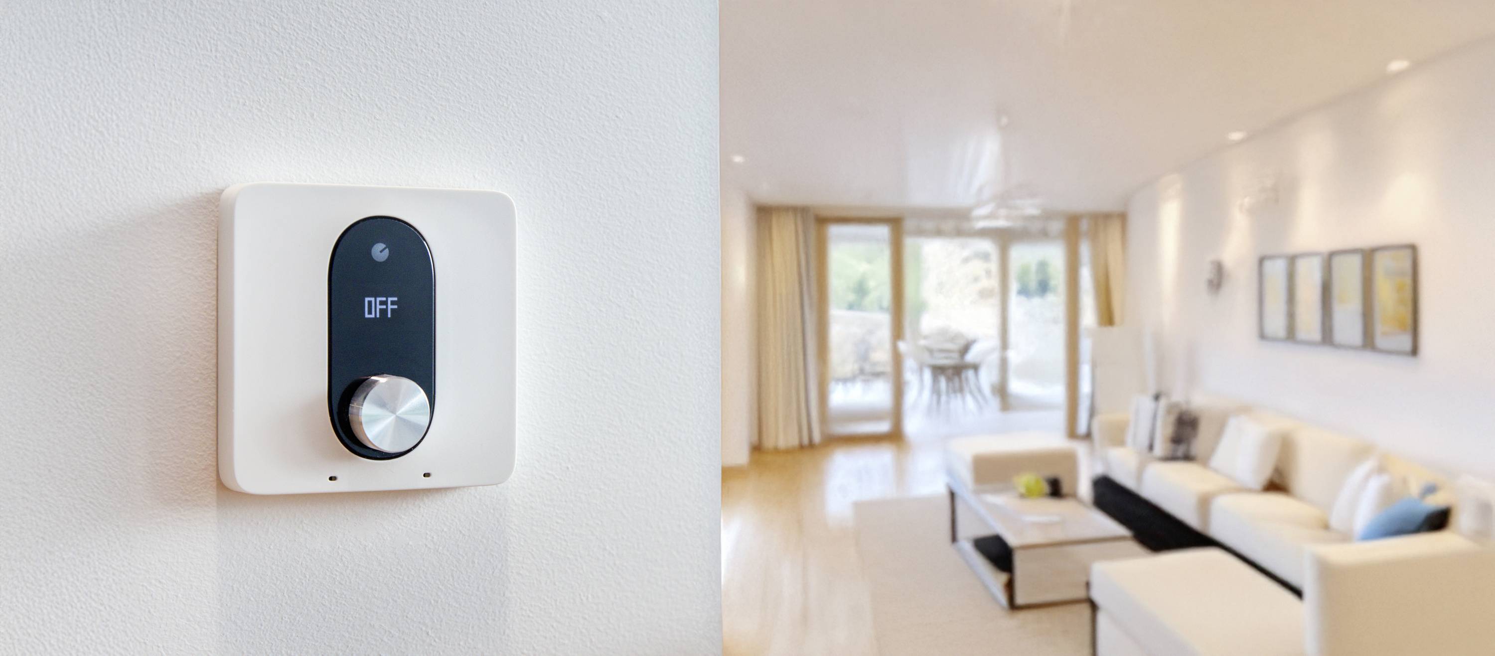 Ein Smart-Thermostat an einer Wand zeigt den Status 'OFF'. Im Hintergrund ein modernes Wohnzimmer mit weißen Möbeln und großen Fenstern.
