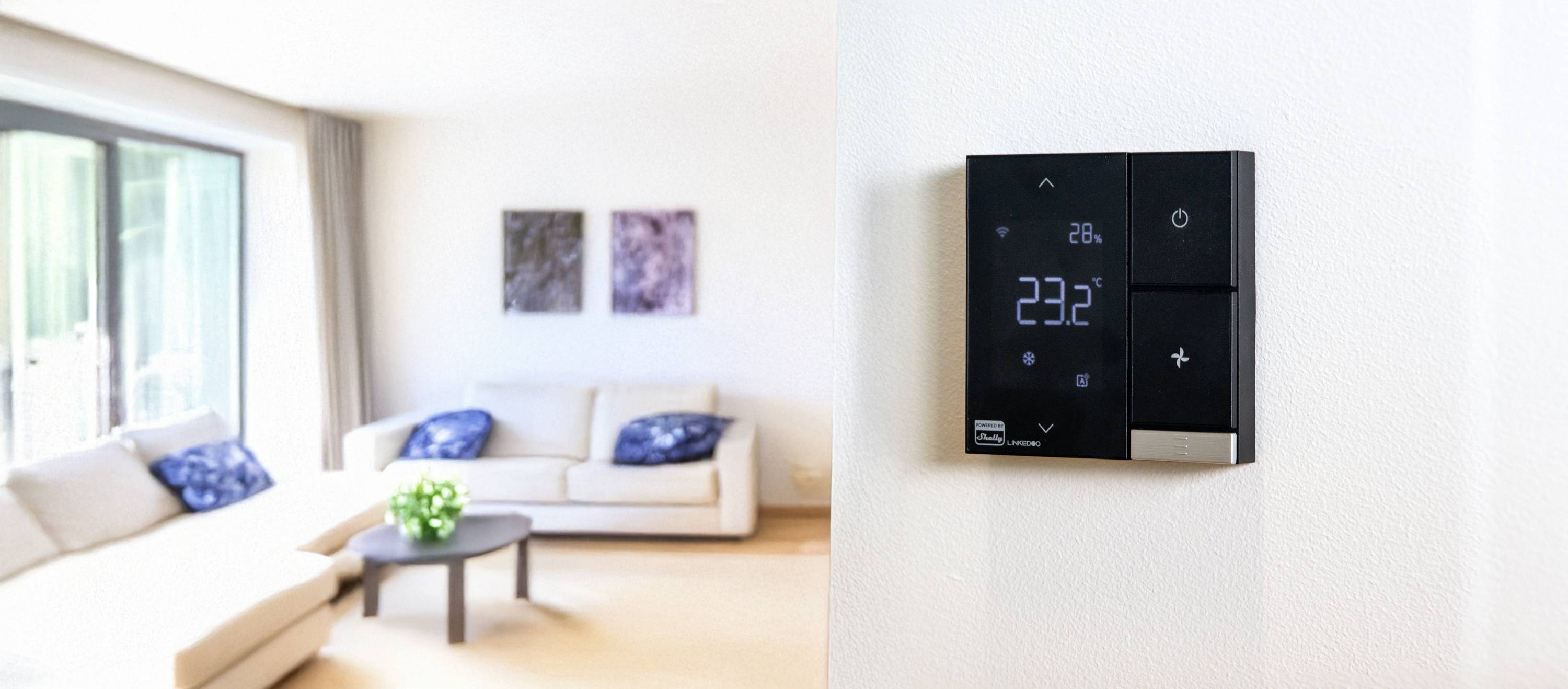Ein modernes Wohnzimmer mit einem digitalen Thermostat an der Wand, das 23,2 Grad anzeigt. Helle Möbel und Kunstwerke im Hintergrund.