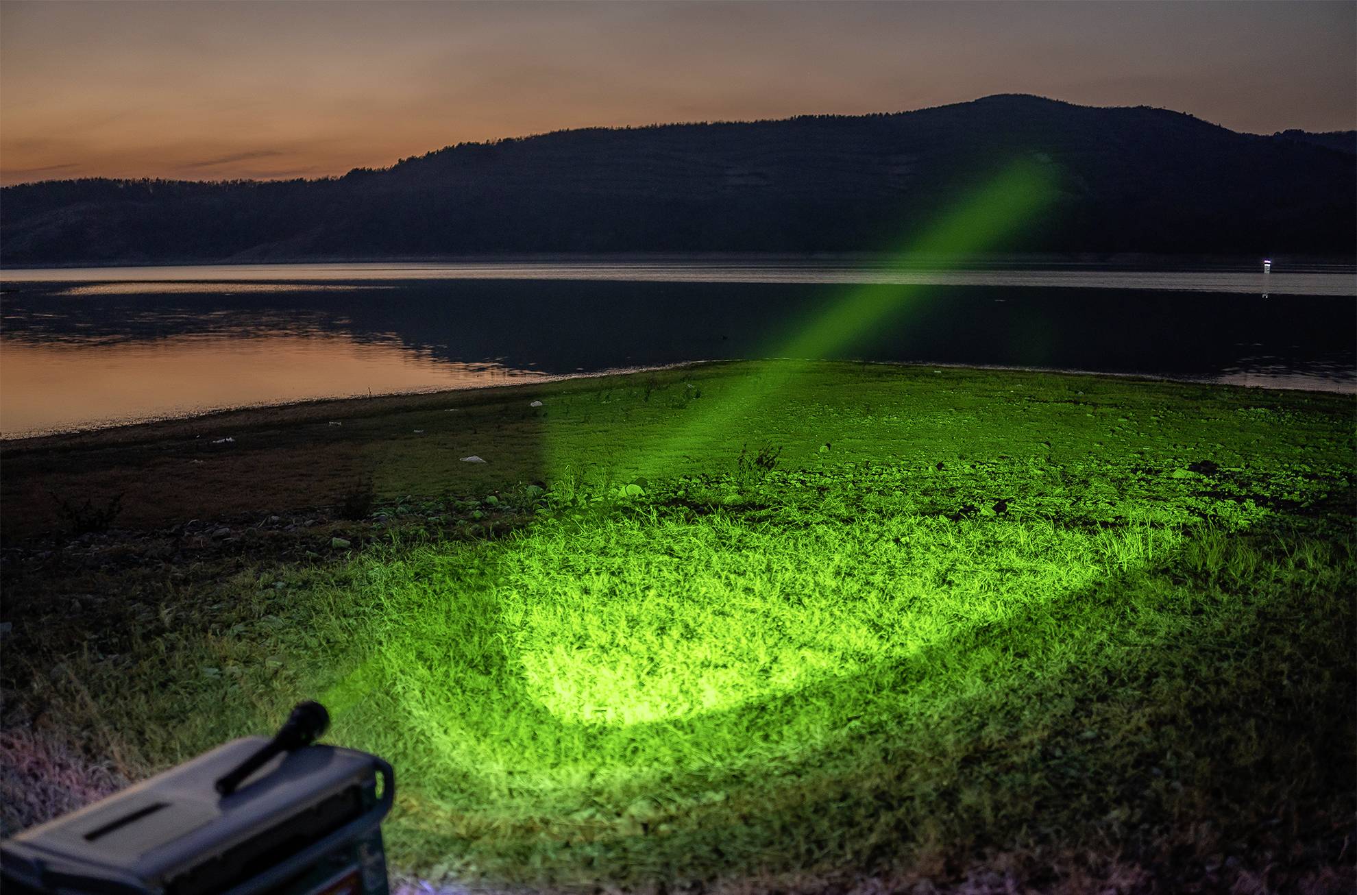 Ein grüner Lichtstrahl strahlt auf eine Wiese am Ufer eines Sees, umgeben von Bergen im Hintergrund bei Dämmerung.