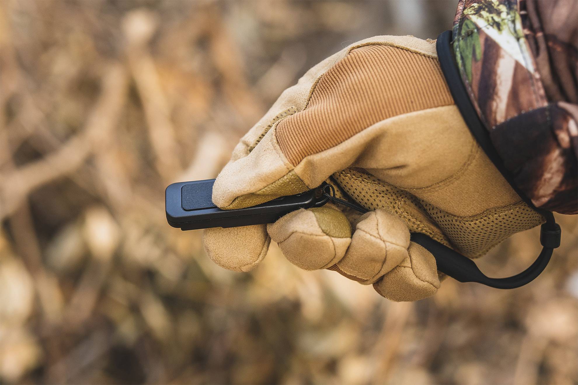 Eine behandschuhte Hand hält ein schwarzes elektronisches Gerät in einem herbstlichen Waldhintergrund.