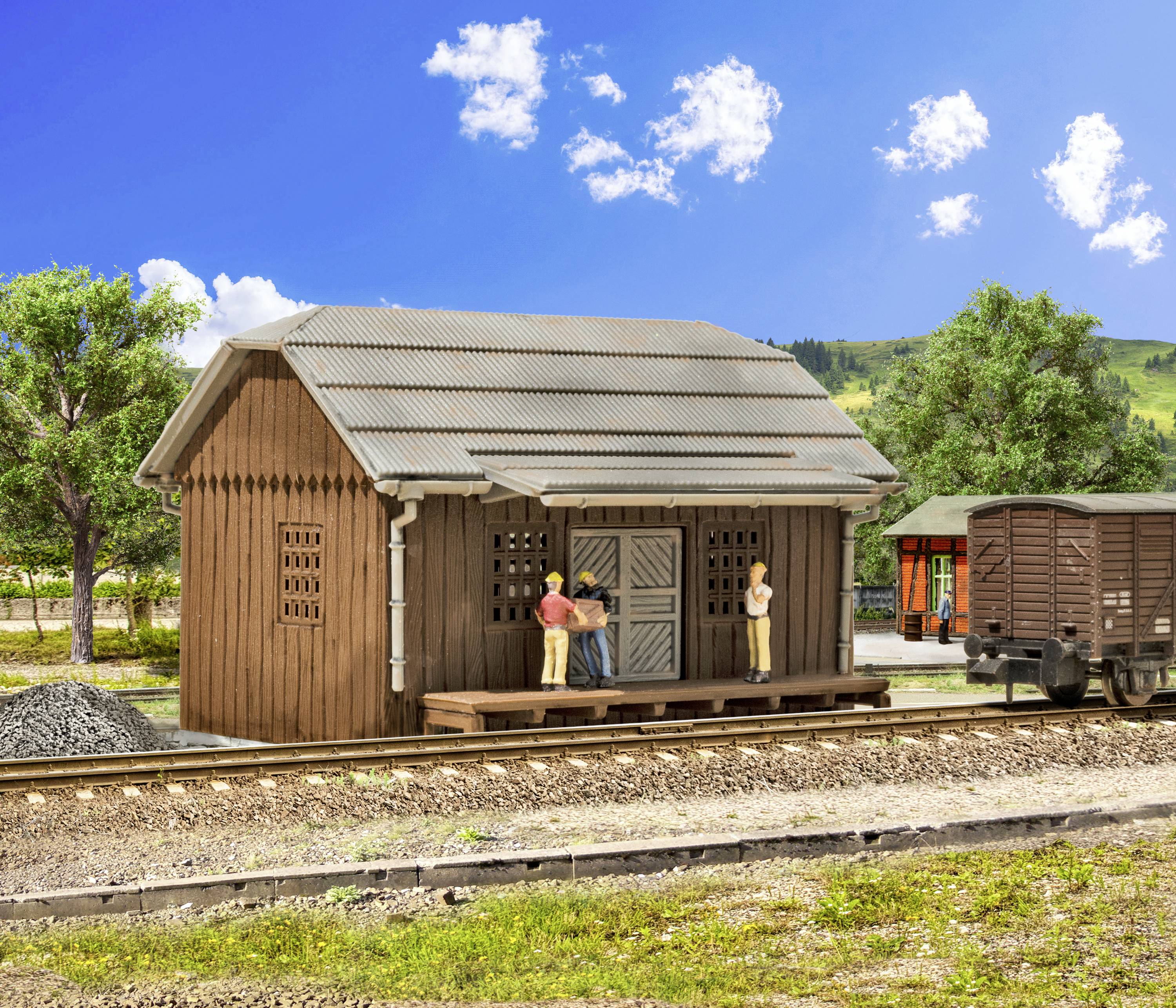 Kleines Holzgebäude an einem ländlichen Bahnsteig, mit drei Figuren, die sich unterhalten. Eisenbahnwaggon und grüne Landschaft im Hintergrund.