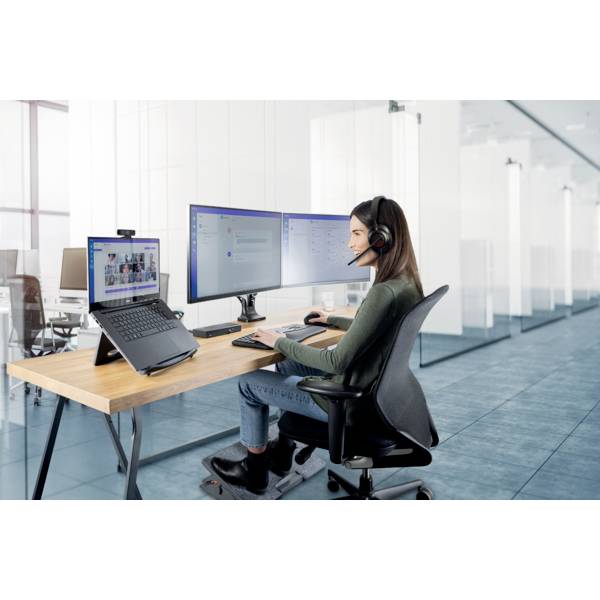 Eine Frau arbeitet an einem Schreibtisch mit zwei Monitoren und Laptop, trägt ein Headset, in einem modernen Büro mit hellen Wänden.