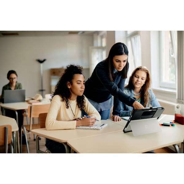 Drei Personen sitzen an einem Tisch und verwenden ein Tablet. Eine Person steht und zeigt auf den Bildschirm. Im Hintergrund arbeitet eine Person an einem Laptop.