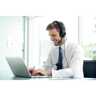 Ein lächelnder Mann trägt ein Headset und arbeitet an einem Laptop in einem modernen Büro. Im Hintergrund sind Fenster zu sehen.