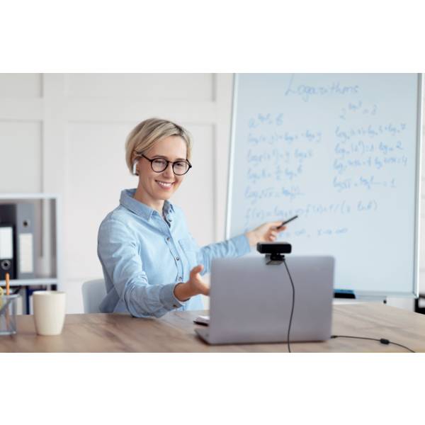 Eine lächelnde Frau in einem blauen Hemd sitzt am Schreibtisch, spricht online und zeigt auf eine Tafel mit mathematischen Formeln.