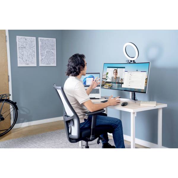 Ein Mann sitzt an einem Schreibtisch und hält eine Videokonferenz auf einem Computerbildschirm. Ein Ringlicht ist über dem Monitor angebracht. Im Hintergrund sind Karten an der Wand.