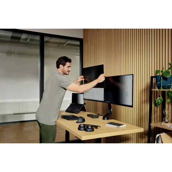 Ein Mann in einem Büro stellt zwei Monitore an einem Schreibtisch ein. Auf dem Tisch liegen verschiedene technische Geräte und Pflanzen.