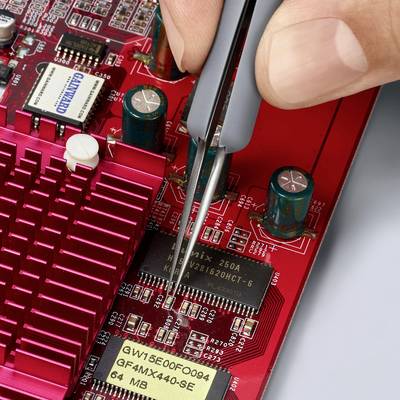 Person hält eine Pinzette, um einen Chip auf einer Computerplatine zu platzieren. Die Platine zeigt mehrere elektronische Bauteile und Beschriftungen.