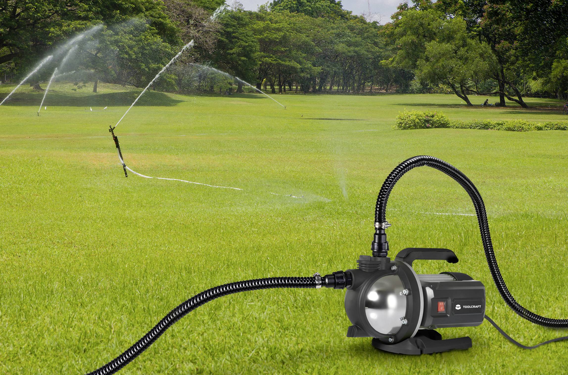 'Gartenbewässerungssystem mit Sprinkler auf einer großen grünen Wiese, im Vordergrund eine schwarze Wasserpumpe, im Hintergrund Bäume.'