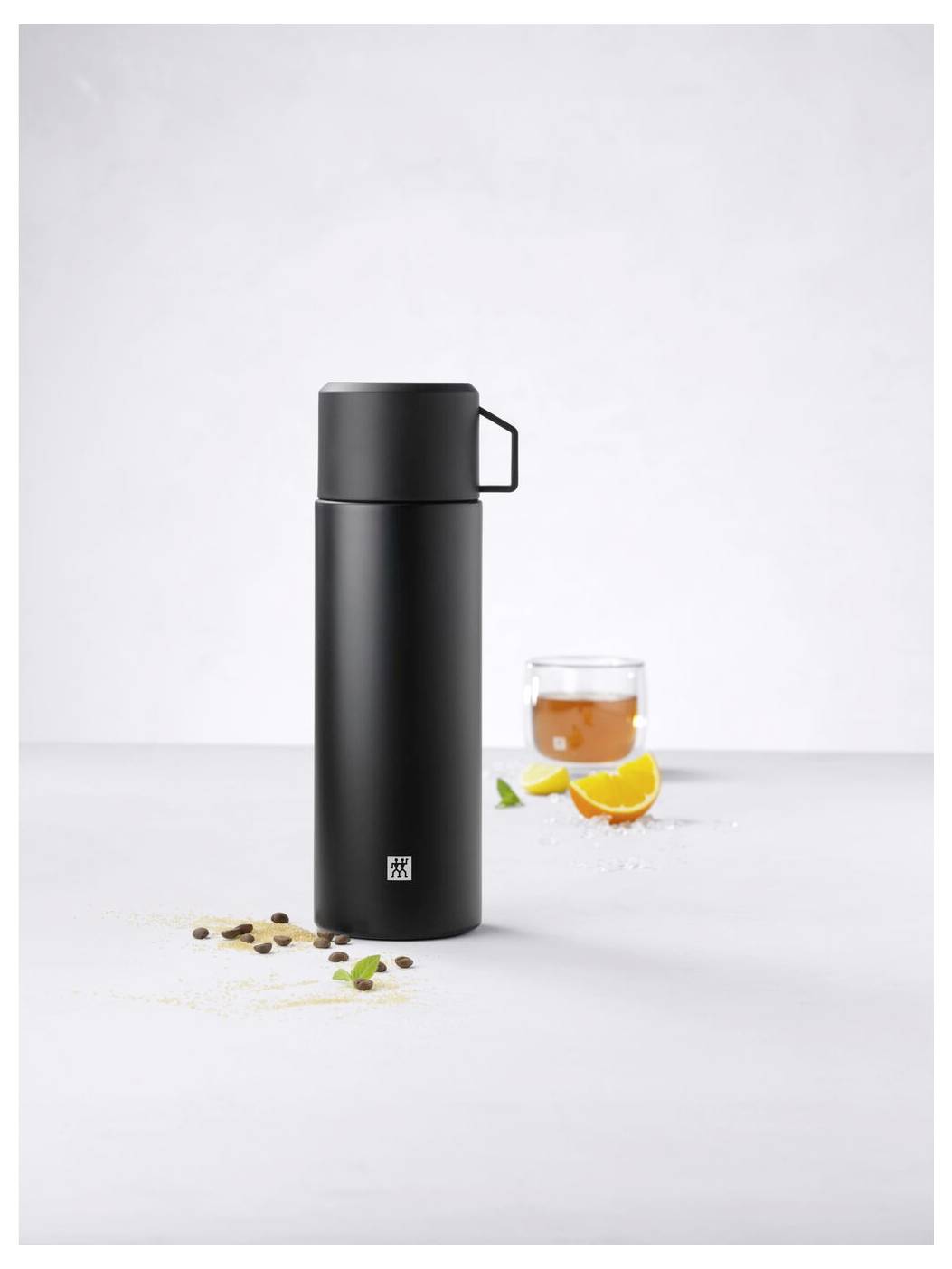 Schwarze Thermosflasche auf weißem Hintergrund, daneben Glas mit Tee und Orangen- und Kräuterdekorationen.